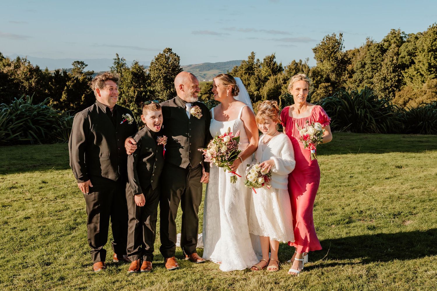 A wedding party poses together on a green hillside during golden hour with mountains in the background.