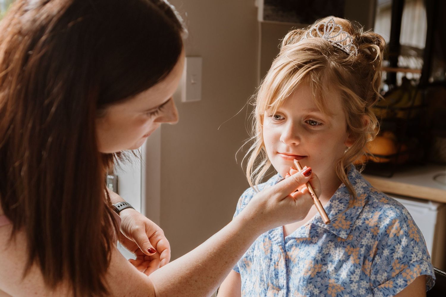 A tender moment as makeup is applied to a small child wearing a light blue patterned shirt in warm indoor lighting.