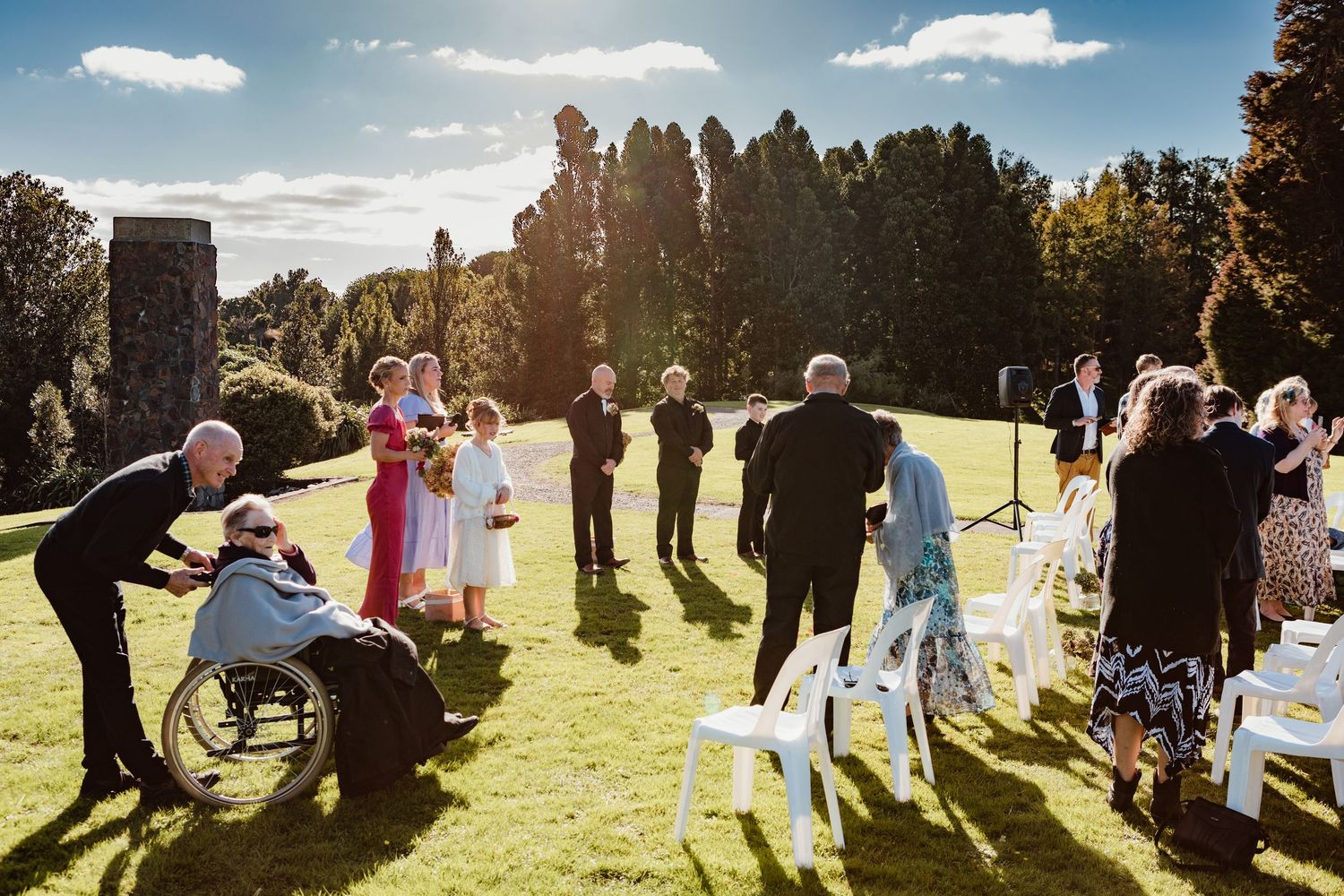 Wedding guests gather on a sunlit lawn with tall trees in the background during an outdoor ceremony.