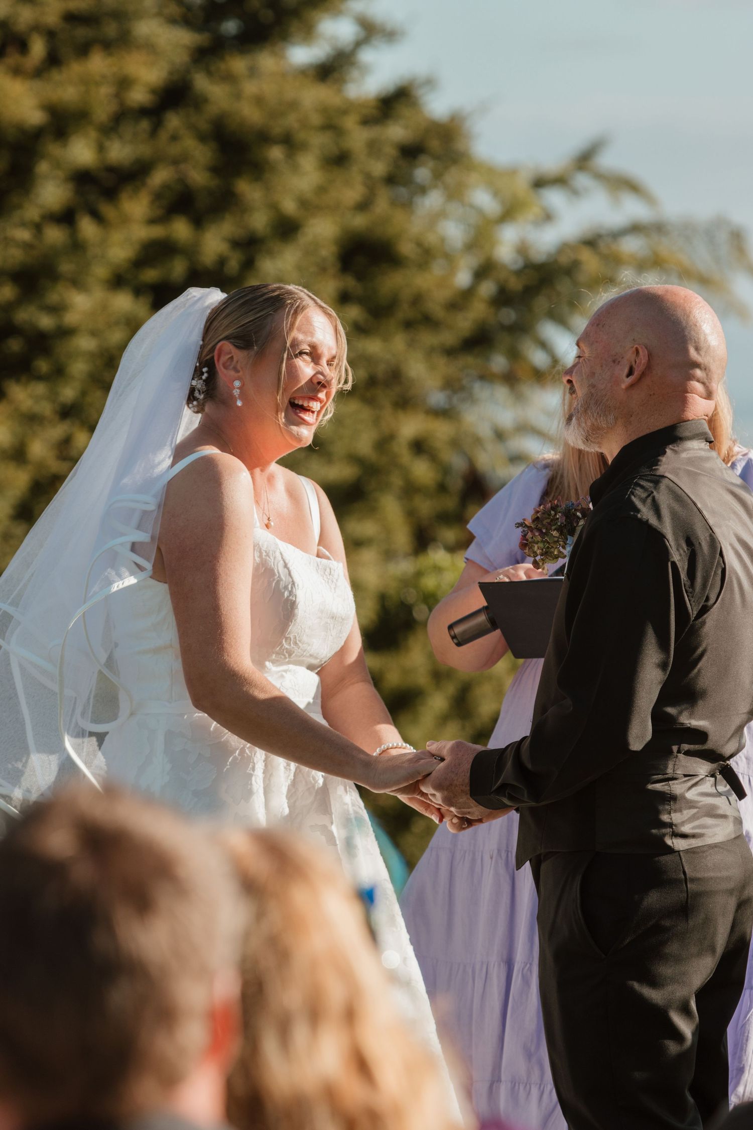 A joyful moment captured during an outdoor wedding ceremony with an officiant and bride in a white dress and veil.