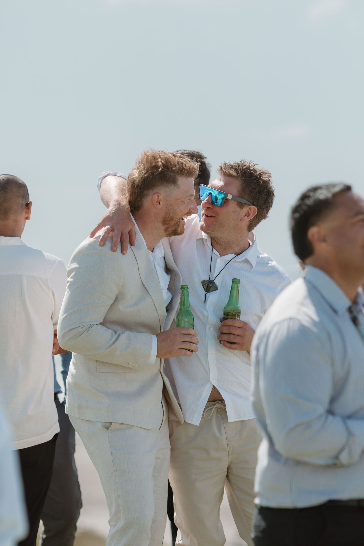 Two men in light suits share a moment holding green beer bottles at an outdoor beach wedding celebration.