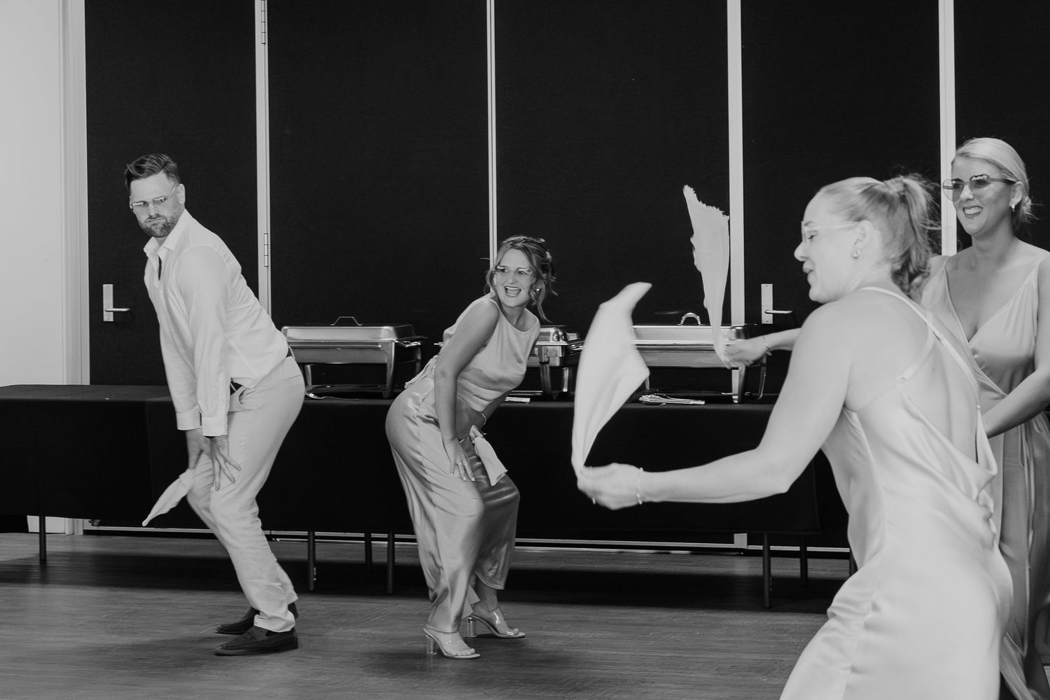 Black and white photo of wedding guests joyfully dancing together in a reception hall.