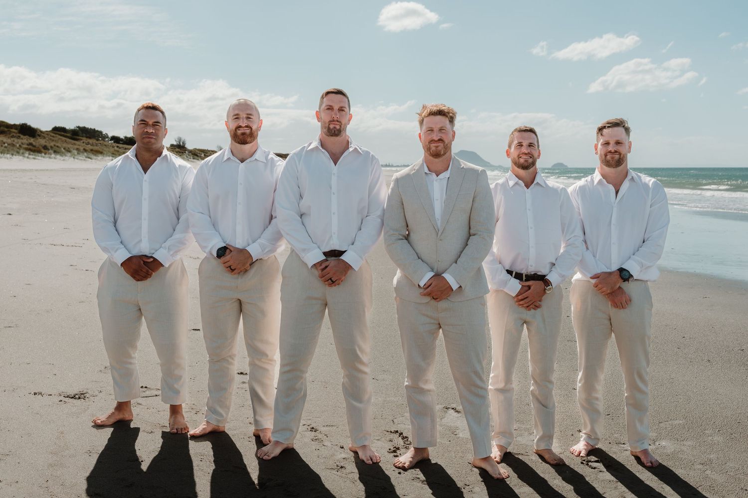 Six groomsmen in white shirts and tan pants pose together on a sunny beach for a wedding photo.