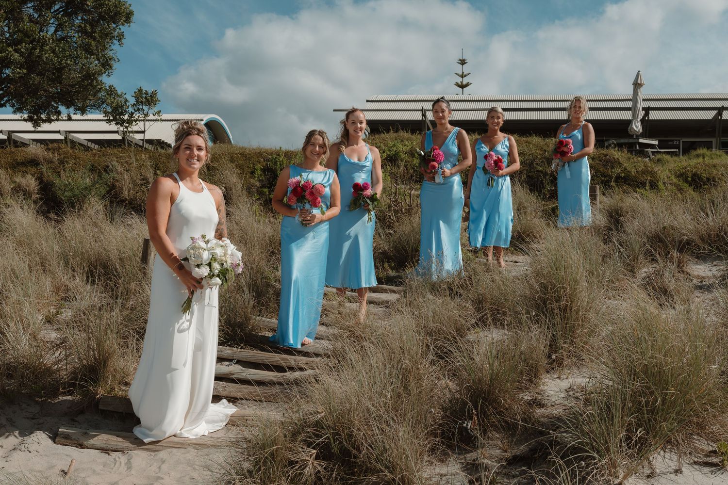 Bride in white gown holds bouquet with bridesmaids in turquoise dresses on sandy beach path.