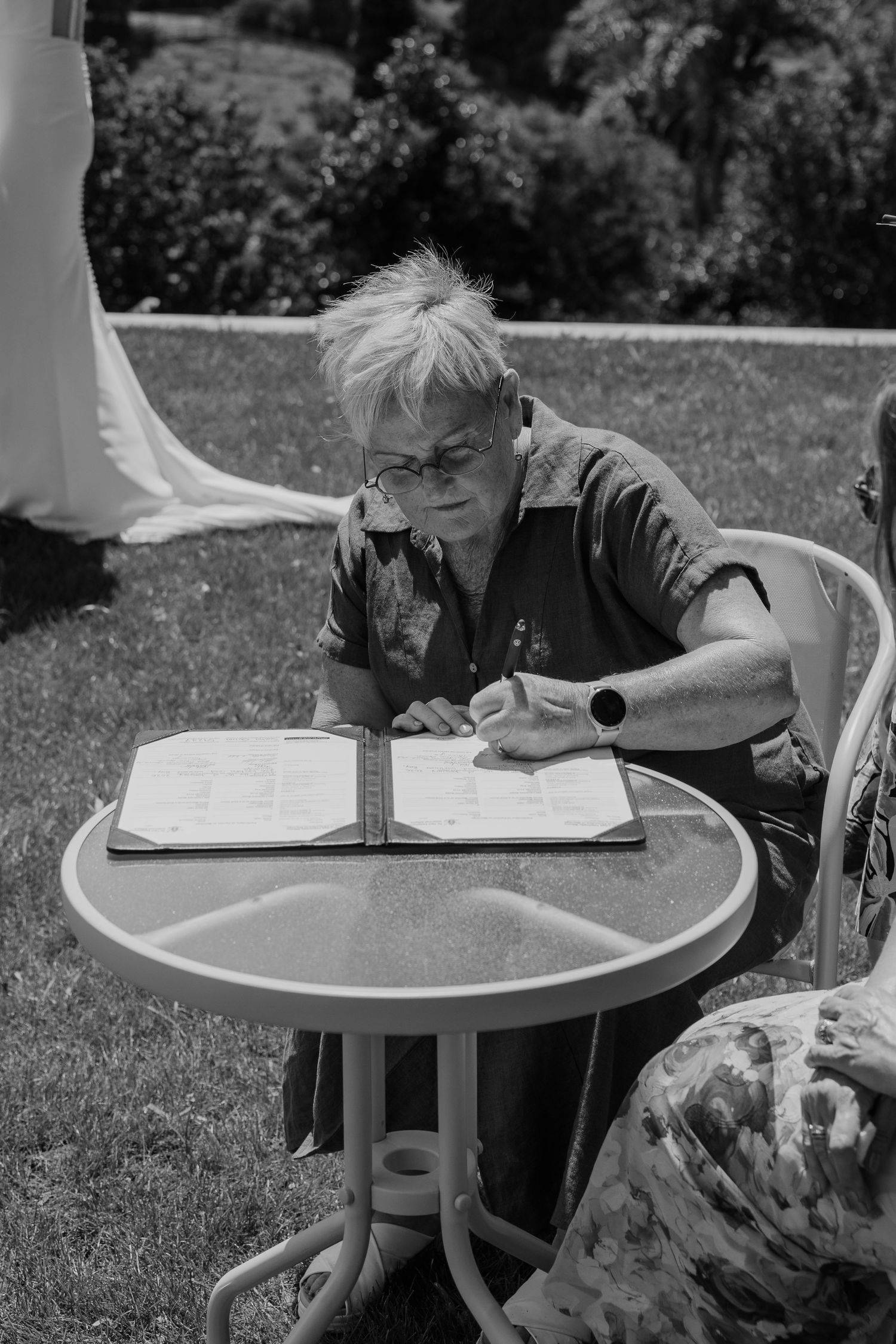 Elderly woman with glasses signing documents at outdoor patio table on sunny day, black and white photo.