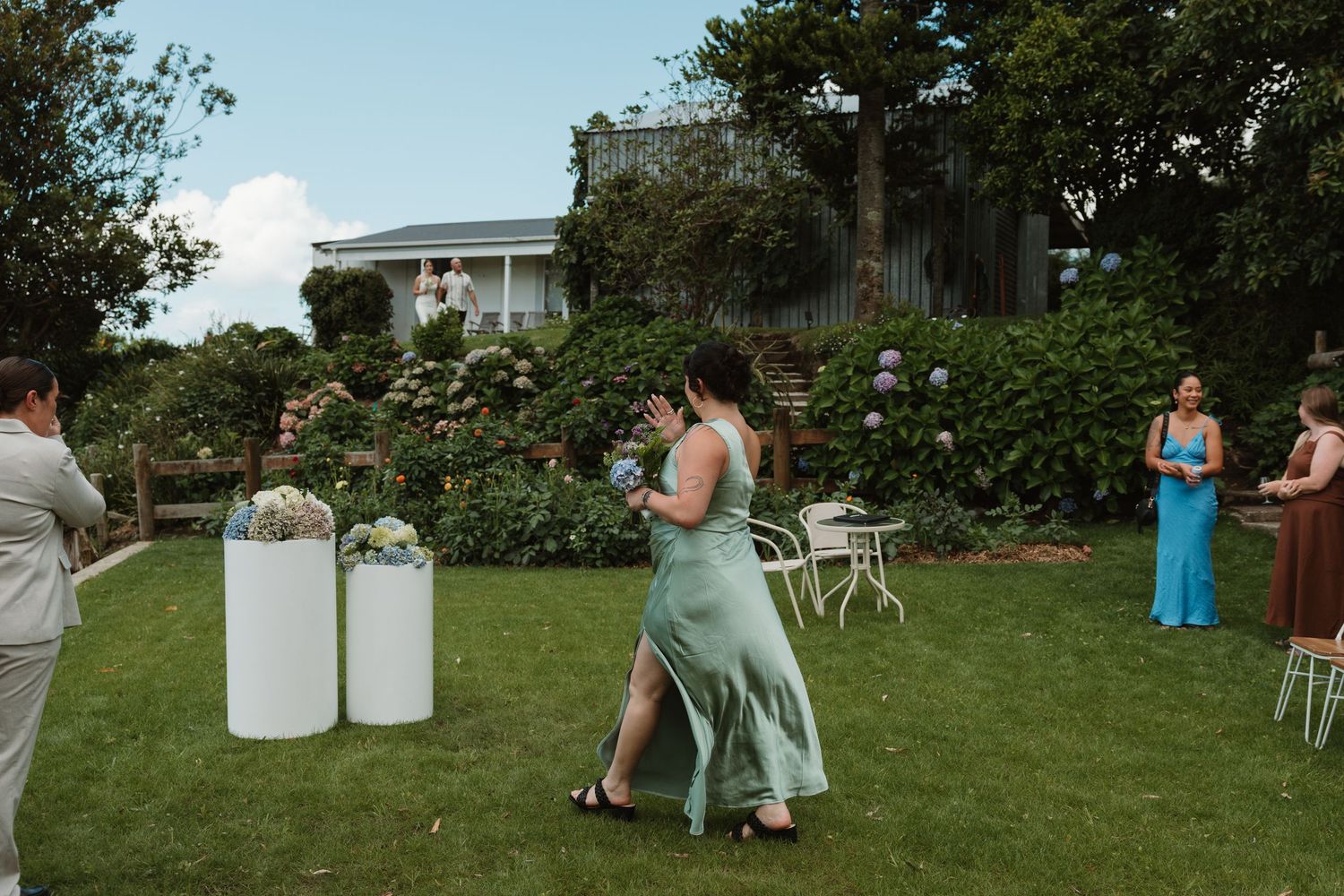 Woman in green dress walks across garden lawn at outdoor wedding ceremony with white pedestals and floral arrangements.
