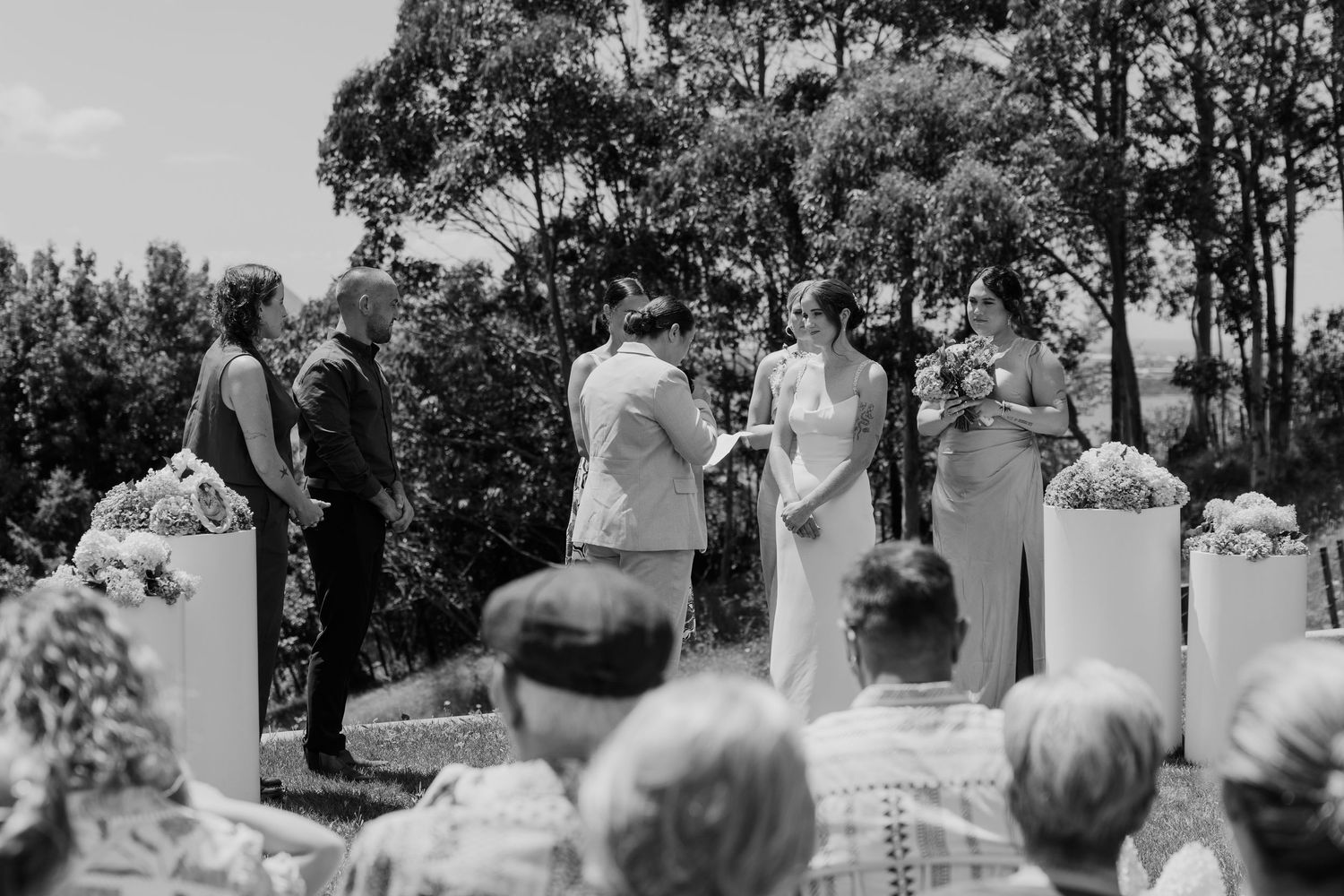 Outdoor wedding ceremony with couple exchanging vows before officiant, guests seated, surrounded by lush greenery.