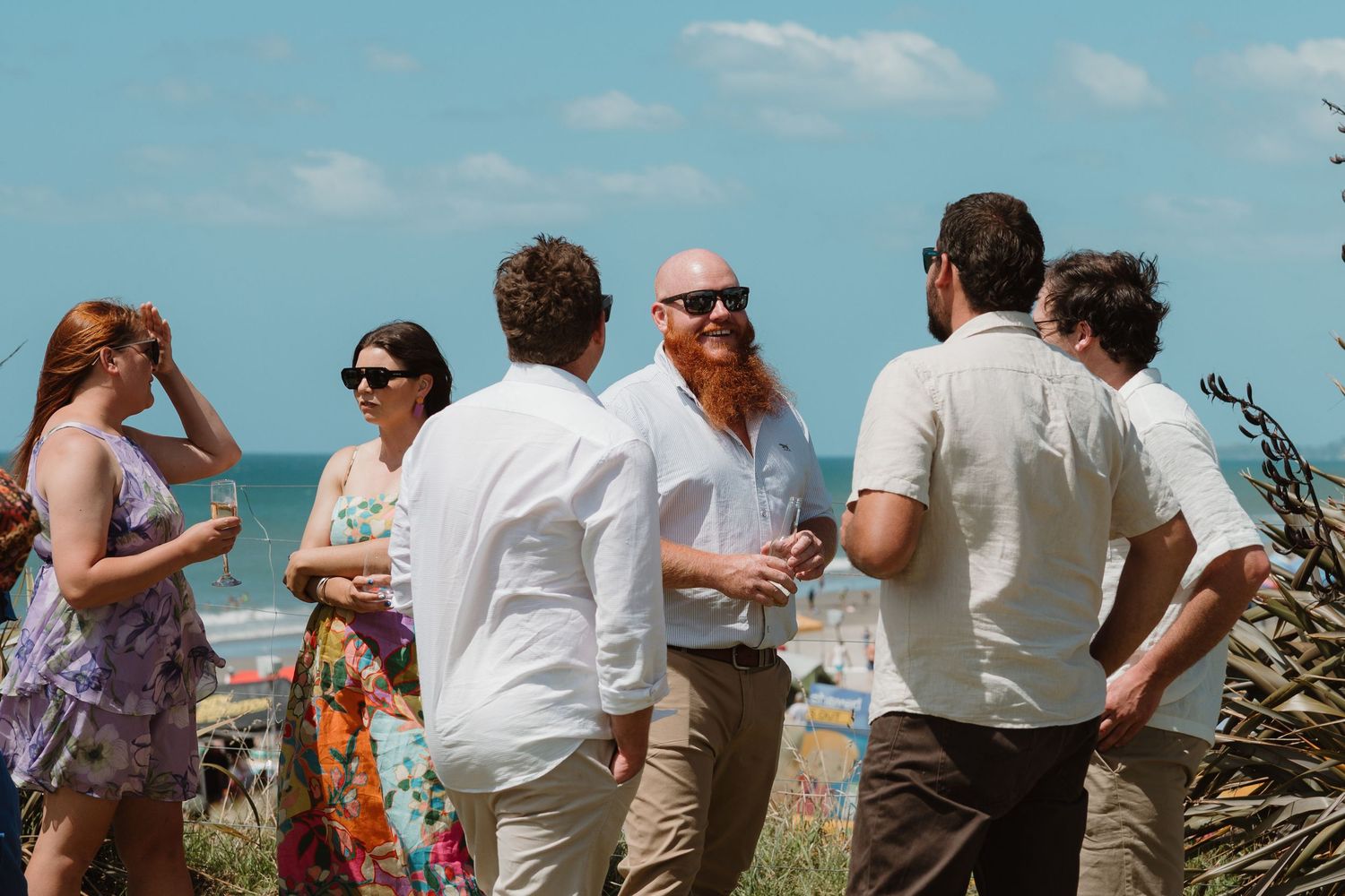 Group of friends socializing at a sunny beach wedding reception with ocean views in the background.