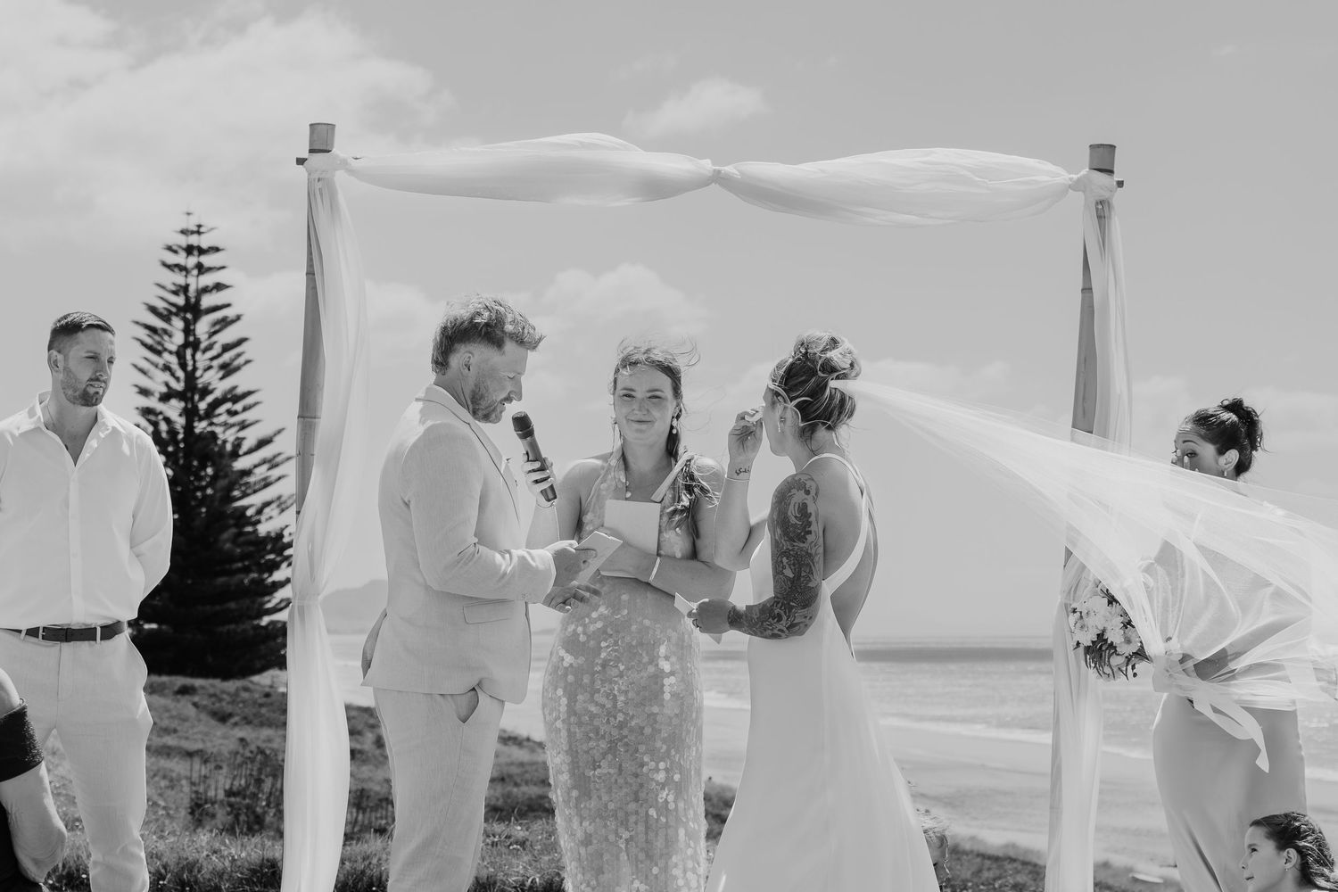 Black and white beach wedding ceremony with couple exchanging vows under a fabric arch, officiant and guests present.
