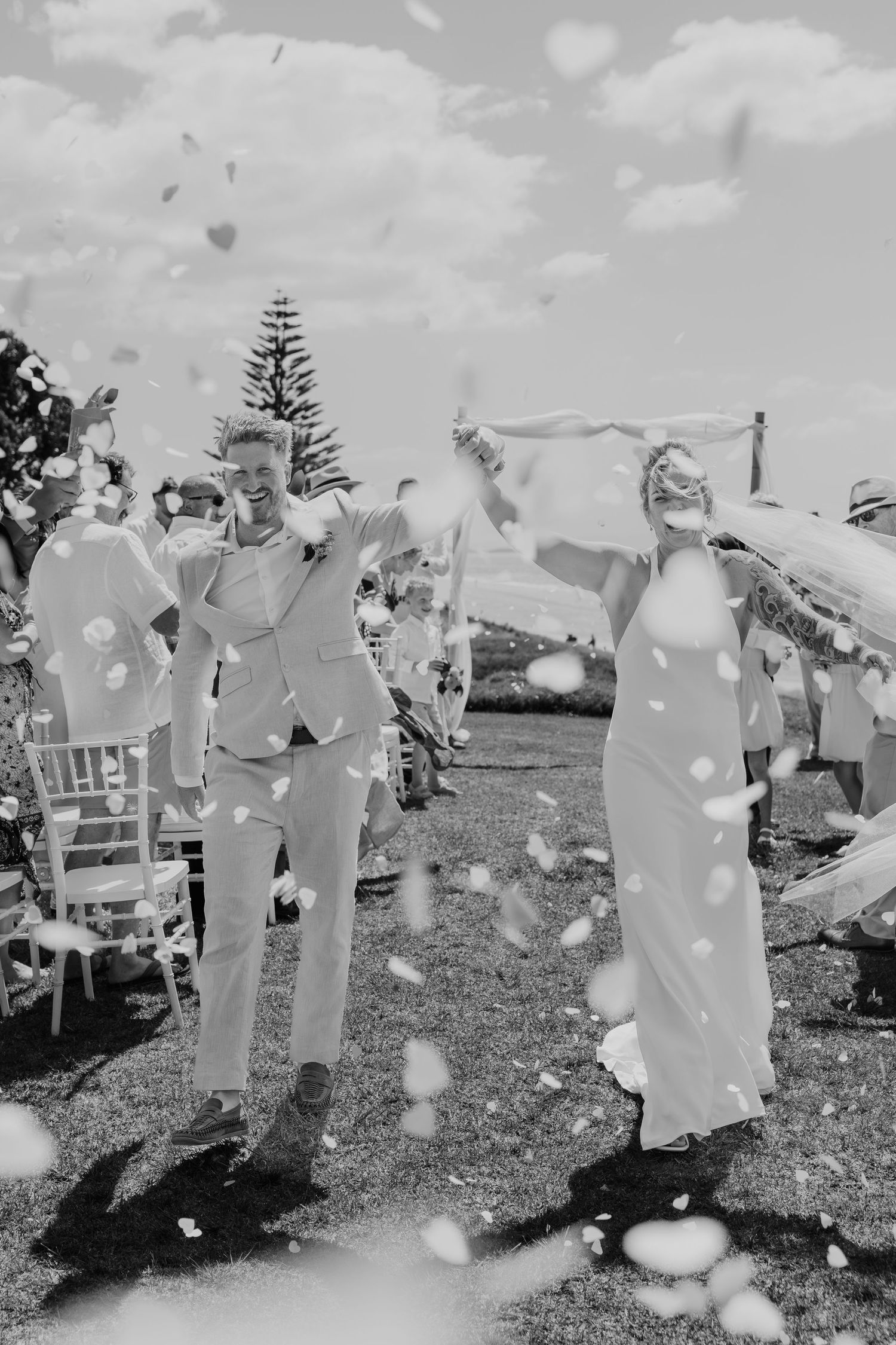 Black and white photo of a joyful couple walking down the aisle with confetti at an outdoor wedding ceremony.