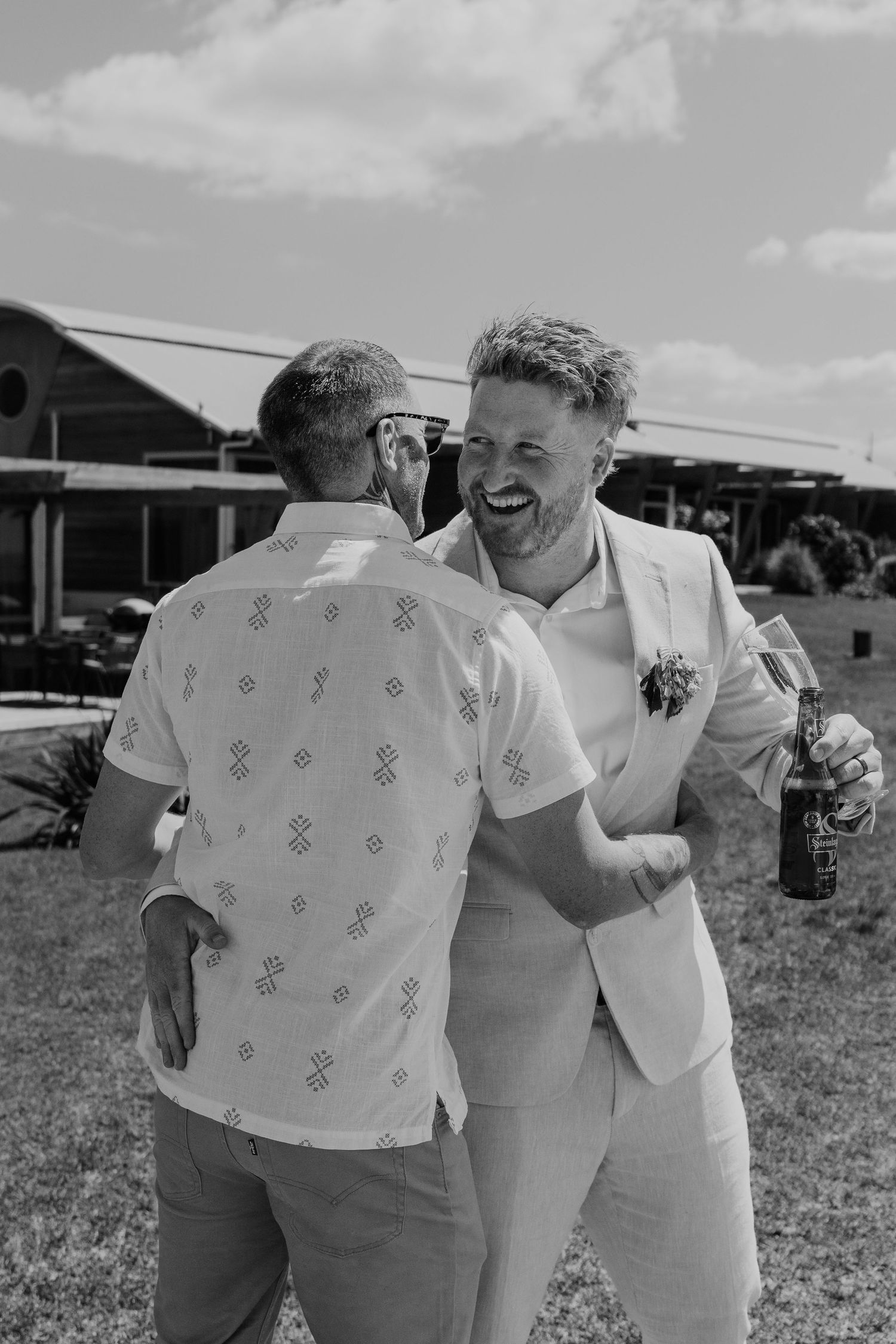 Two men laughing and embracing outdoors at a wedding celebration, one wearing a suit with boutonniere holding a beer bottle.