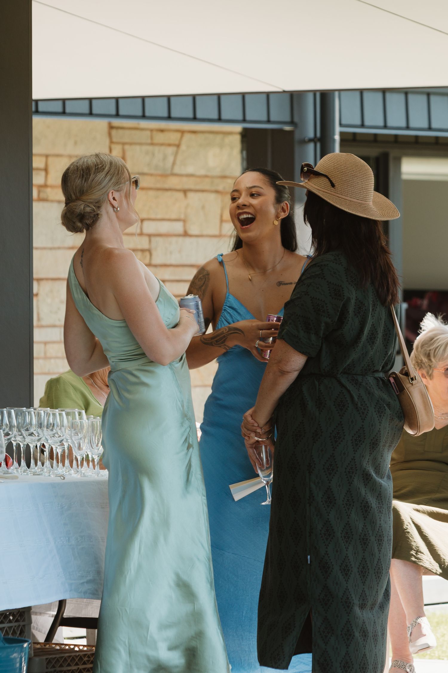 Three women in elegant dresses laugh together at an outdoor event, holding drinks near a table of champagne glasses.