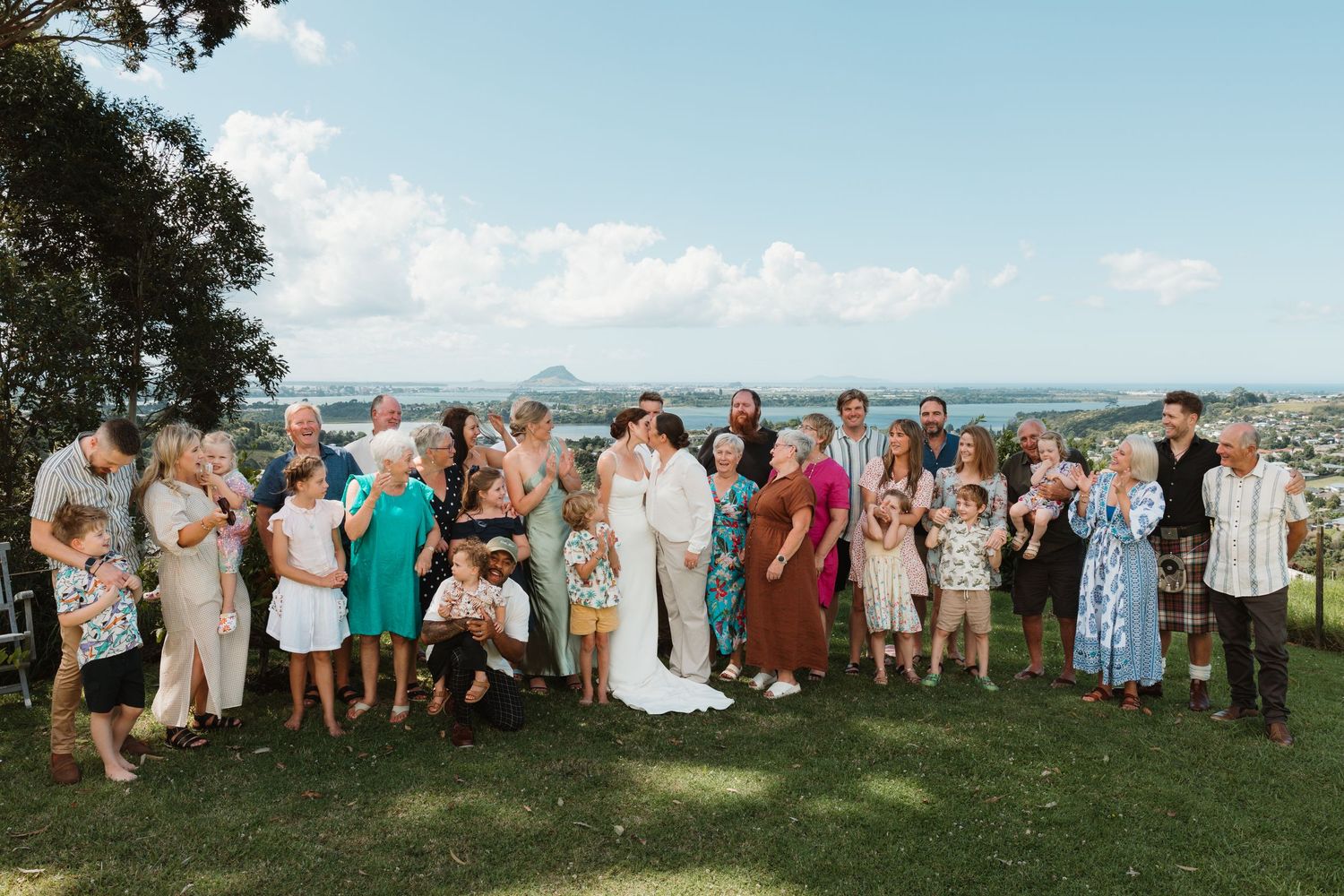 Large wedding group photo outdoors with ocean views, bride and groom surrounded by family and friends on a sunny day.