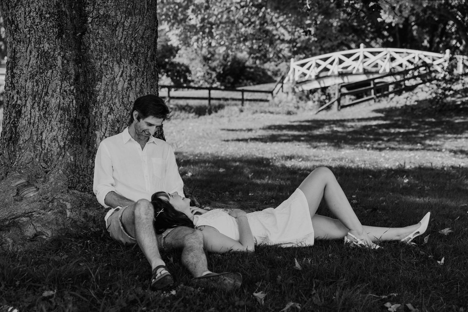 Black and white photo of a couple relaxing under a tree in a park, with a white bridge visible in the background.