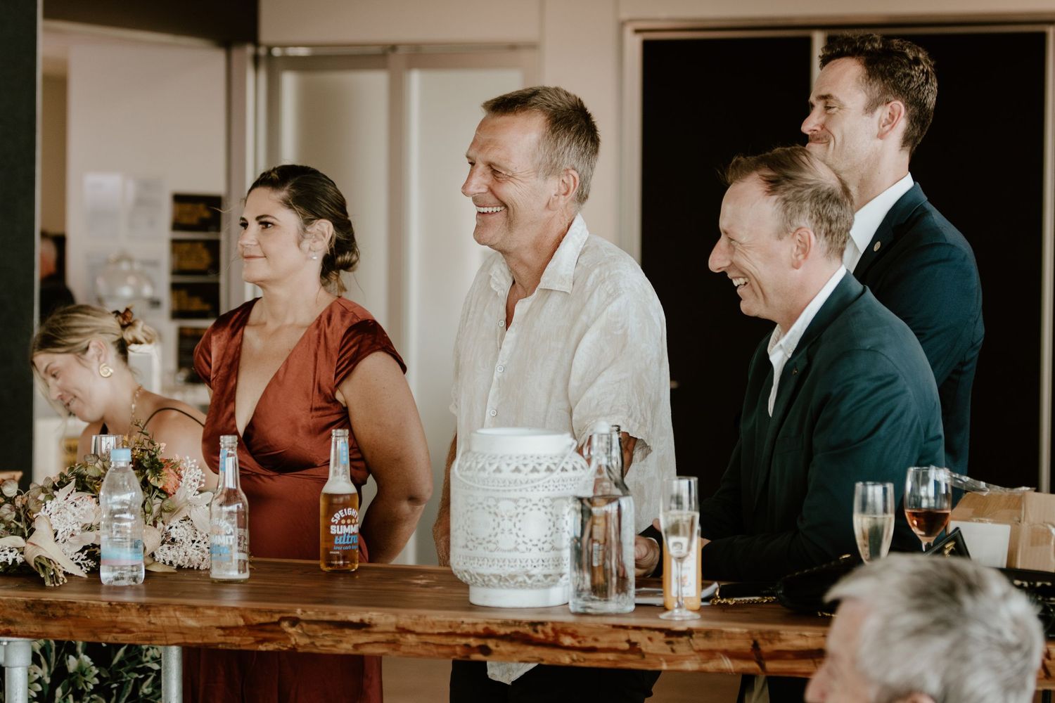A group of people laugh around a wooden table with champagne glasses and a white cake during a celebration.