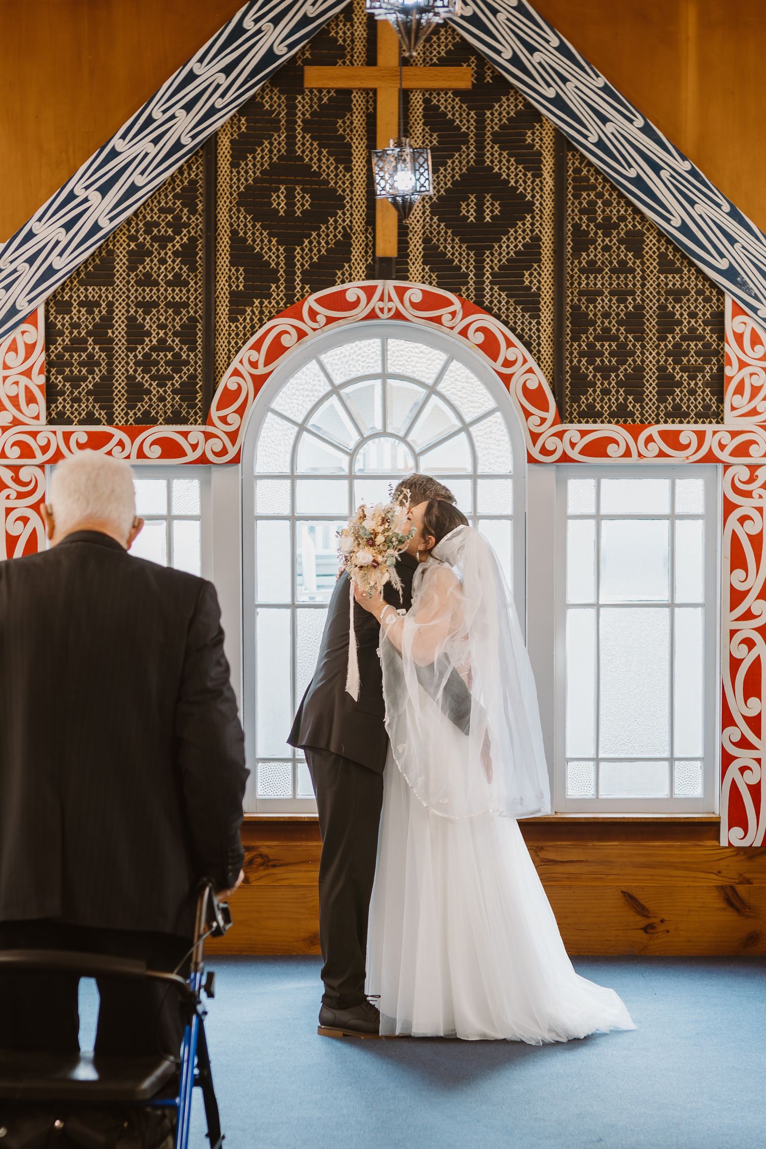 A couple shares their first kiss as newlyweds in a church with ornate wood carvings and a grand arched window.