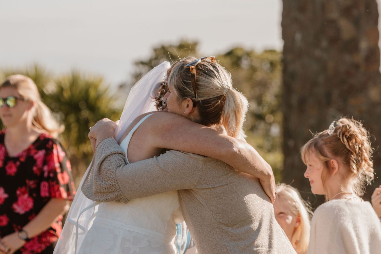 Wedding guests share an emotional embrace during an outdoor ceremony in warm golden sunlight.