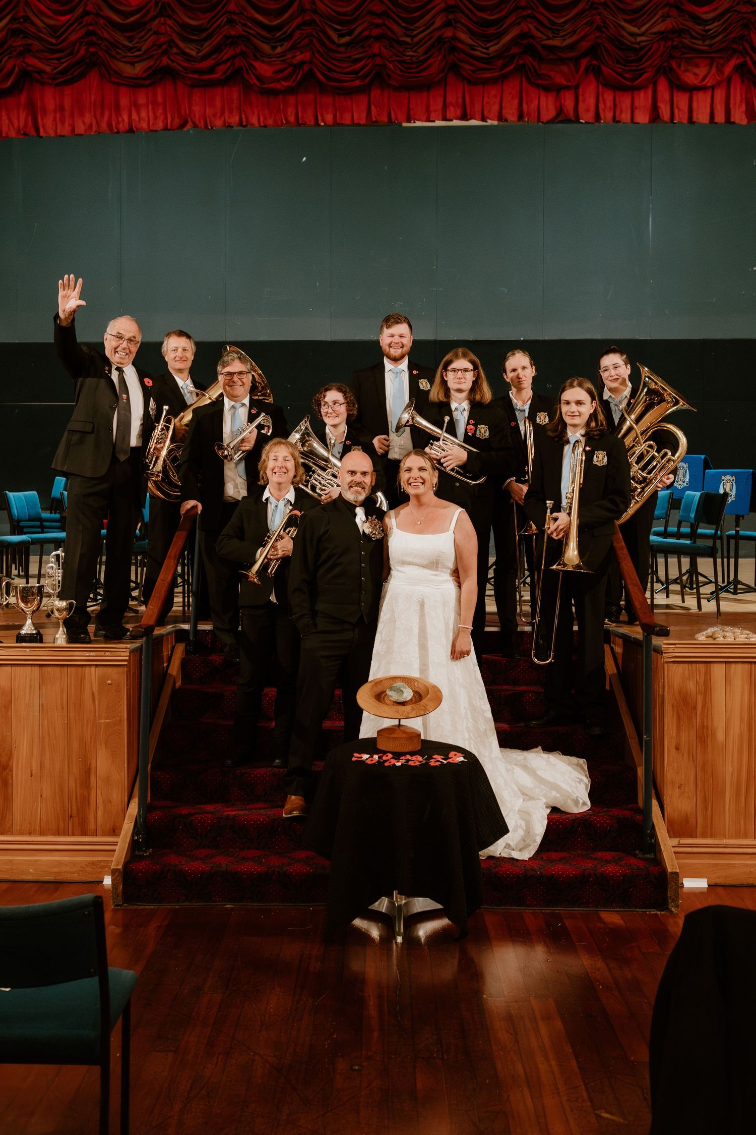 A brass band poses on red-carpeted stairs with their instruments while a person in a white wedding dress sits in front.