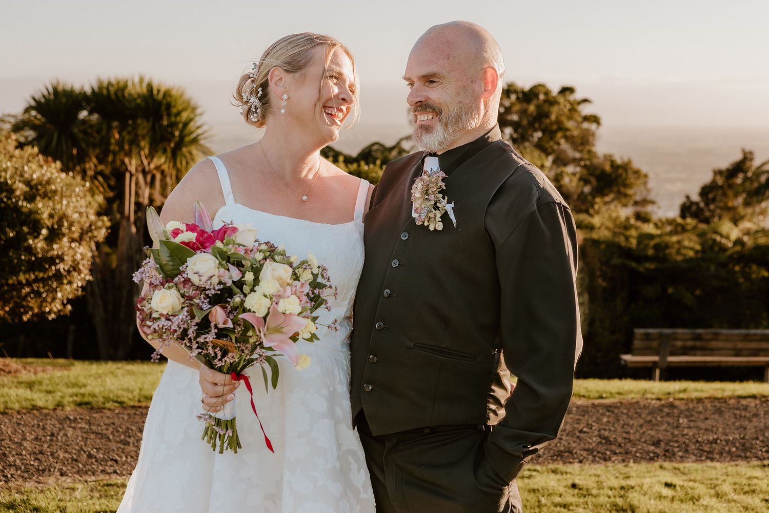 A newly married couple stands together during golden hour in a scenic outdoor setting holding a floral bouquet.
