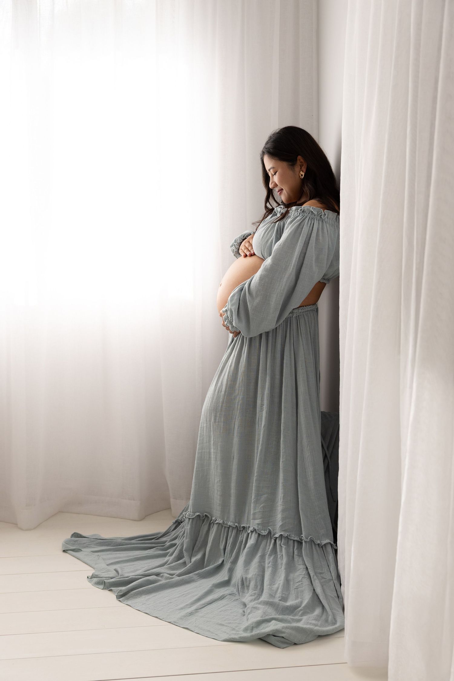 Expecting person wearing a flowing sage green maxi dress stands contemplatively by sheer curtains in natural light.