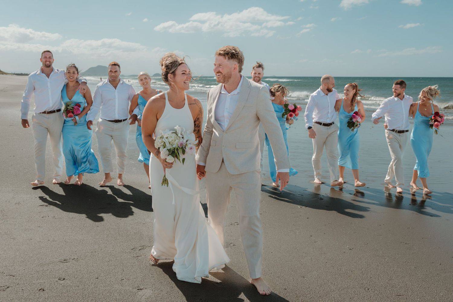 Wedding party walks on beach, bride in white gown, groom in tan suit, bridesmaids in blue dresses, groomsmen in white shirts.
