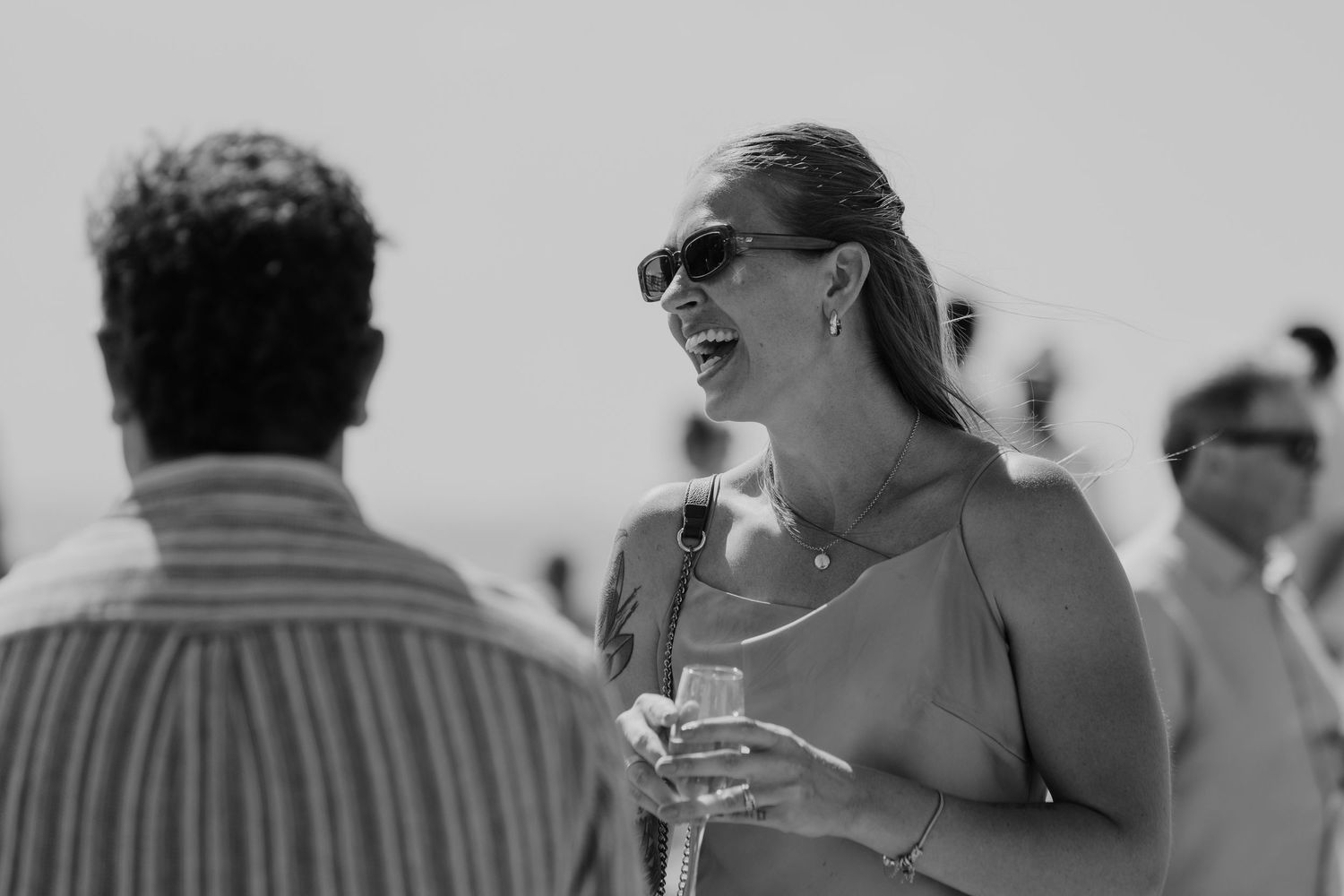 Woman in sunglasses laughing and holding a drink at an outdoor social gathering in black and white photo.
