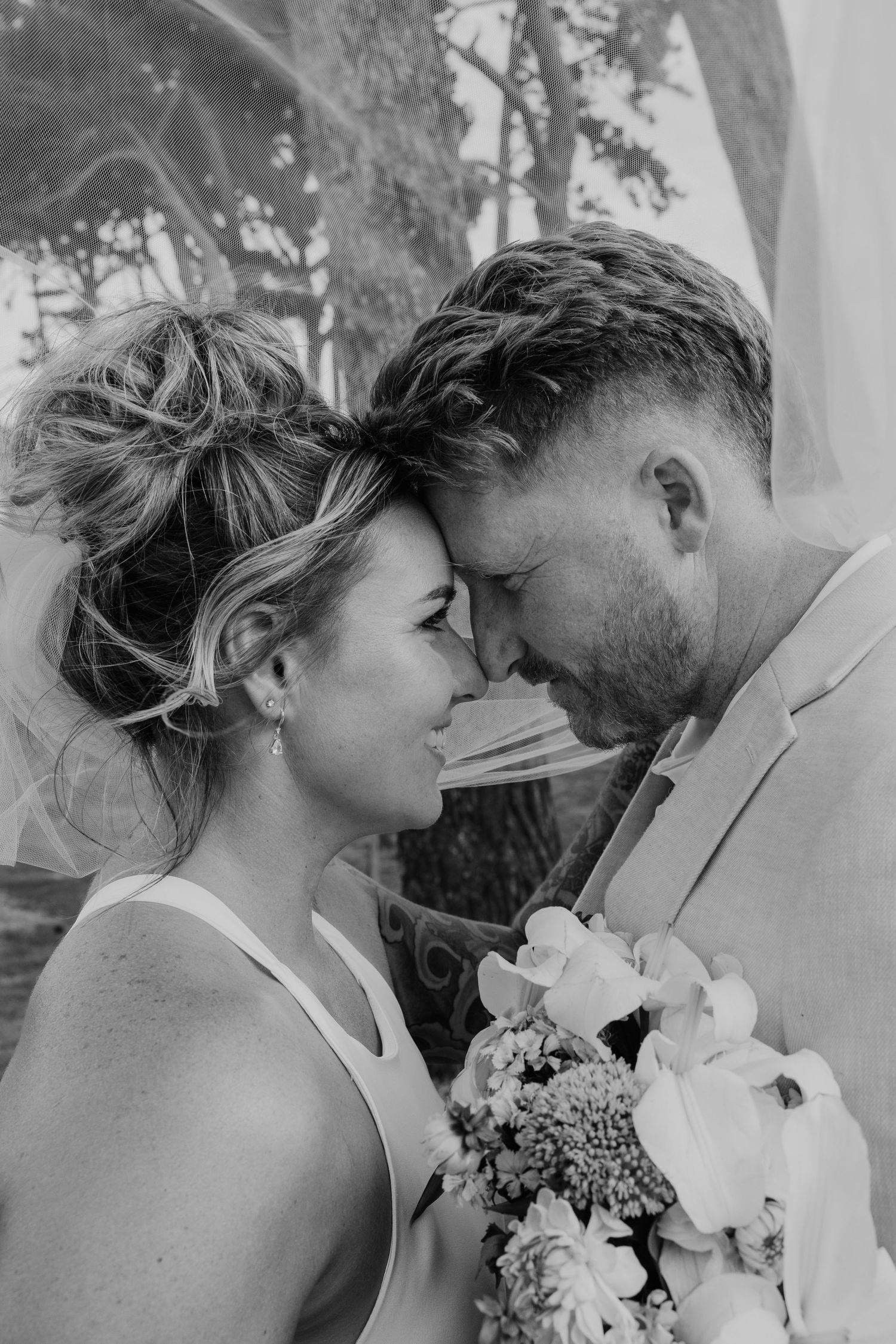 Black and white wedding photo of bride and groom touching foreheads, bride holding floral bouquet outdoors.