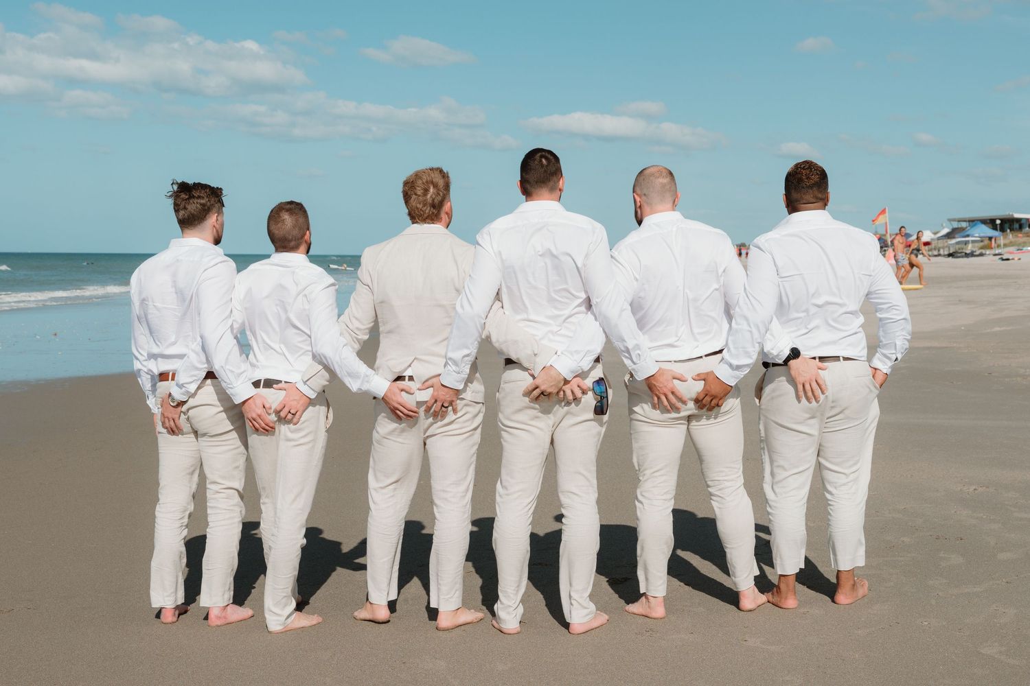 Six groomsmen in white shirts and khaki pants stand barefoot on a sandy beach with their backs to the camera.