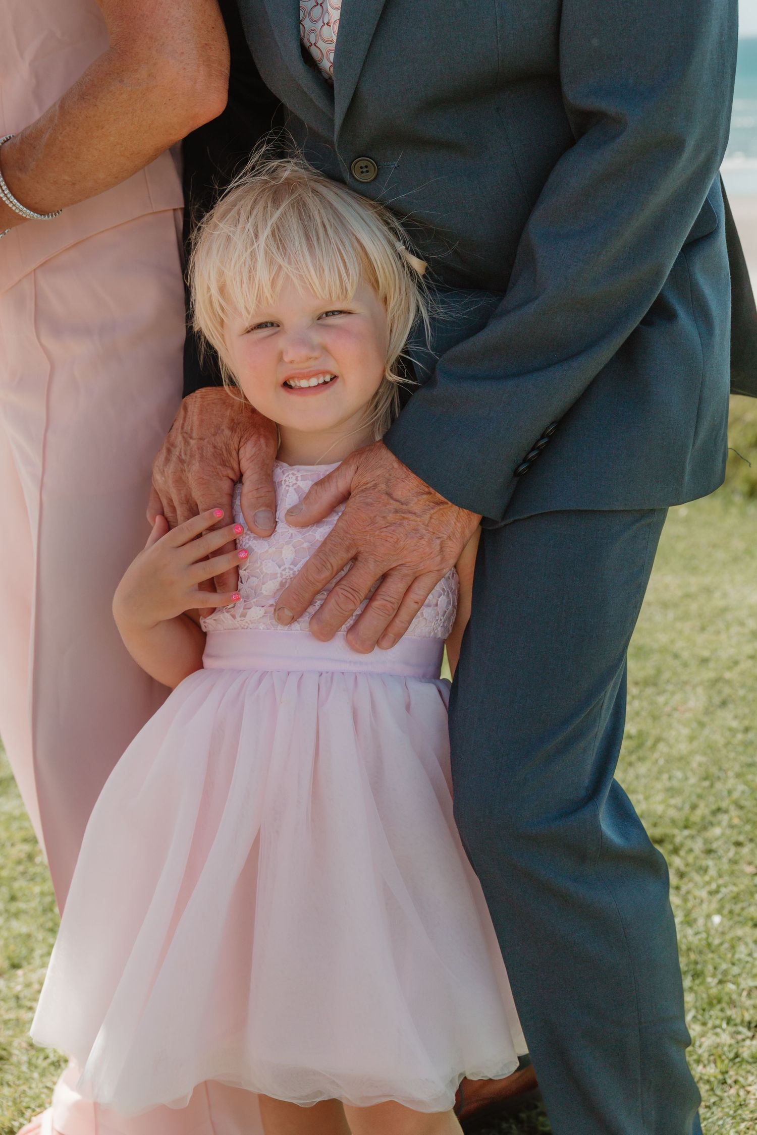 A smiling toddler girl in a pink dress stands between adults in formal attire at an outdoor wedding celebration.