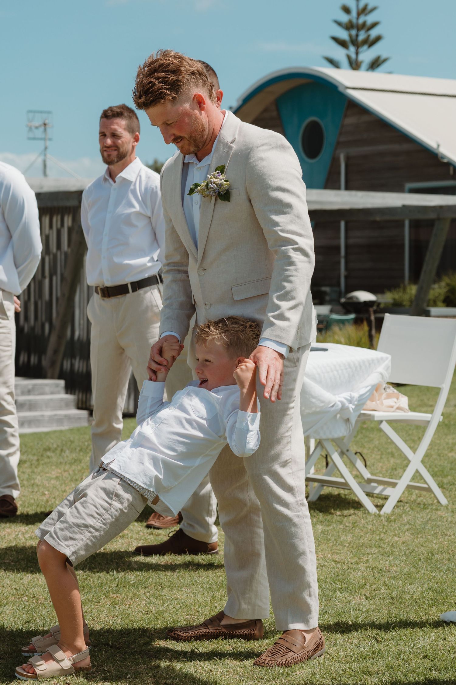 Groom in beige suit playfully holds hands with young ring bearer on lawn at outdoor beach wedding.
