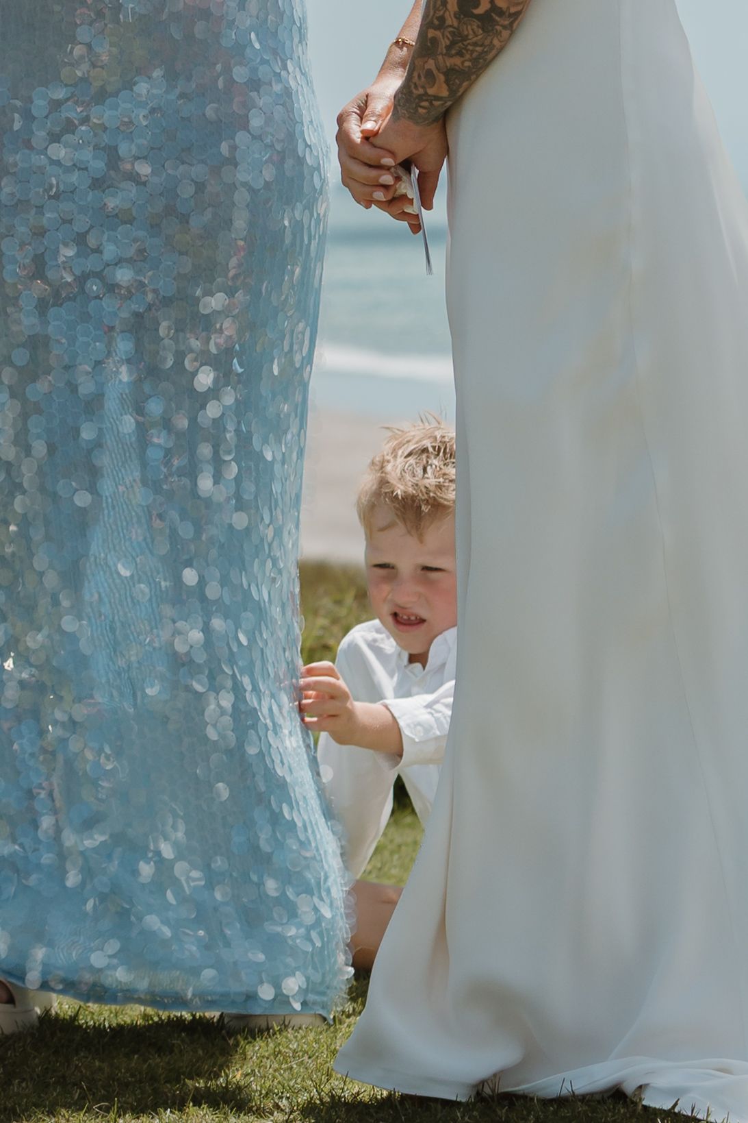 A young boy in white sits between two women in formal dresses at a beach wedding ceremony.