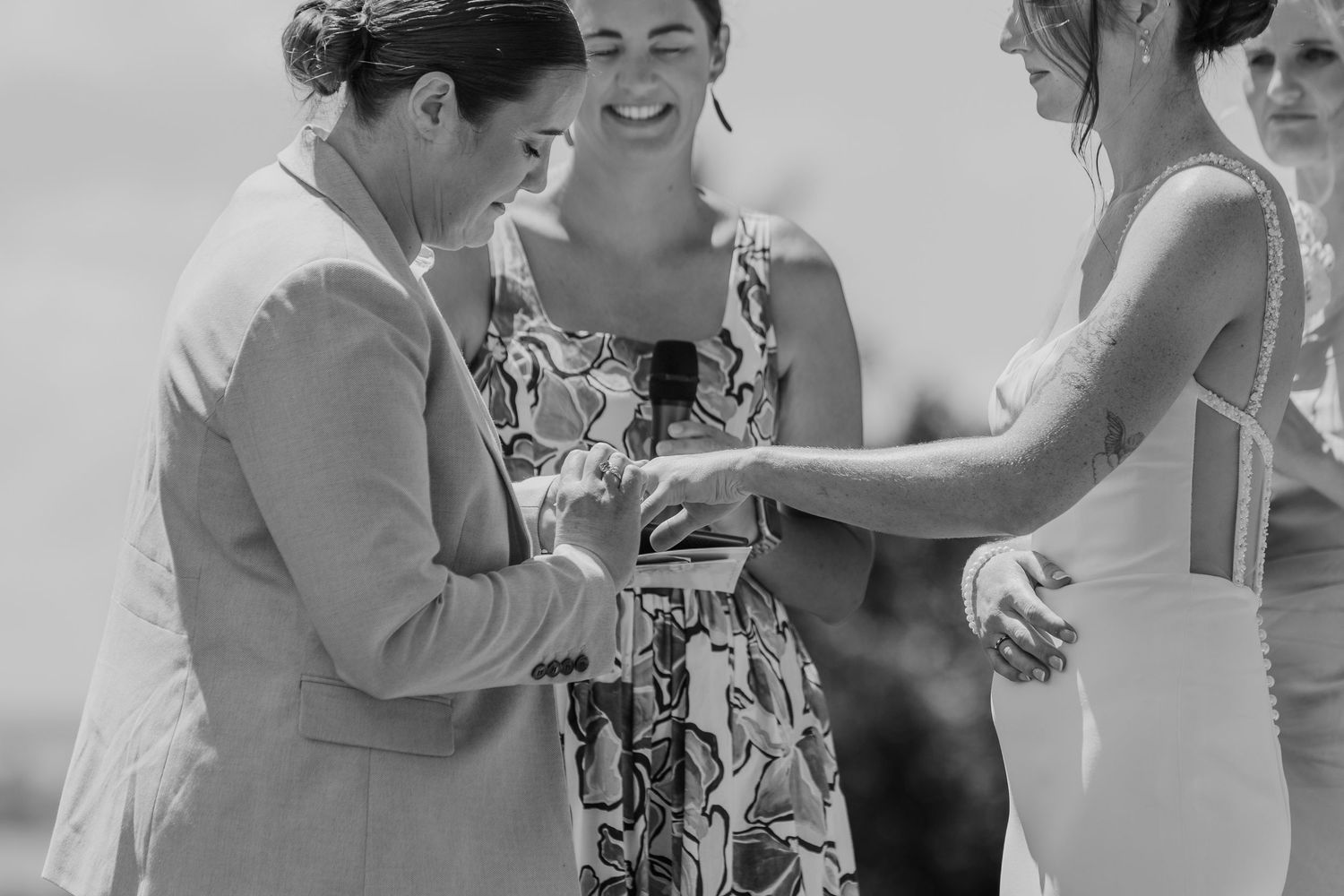 Black and white wedding ceremony photo of two brides exchanging rings while officiant and guest smile joyfully.