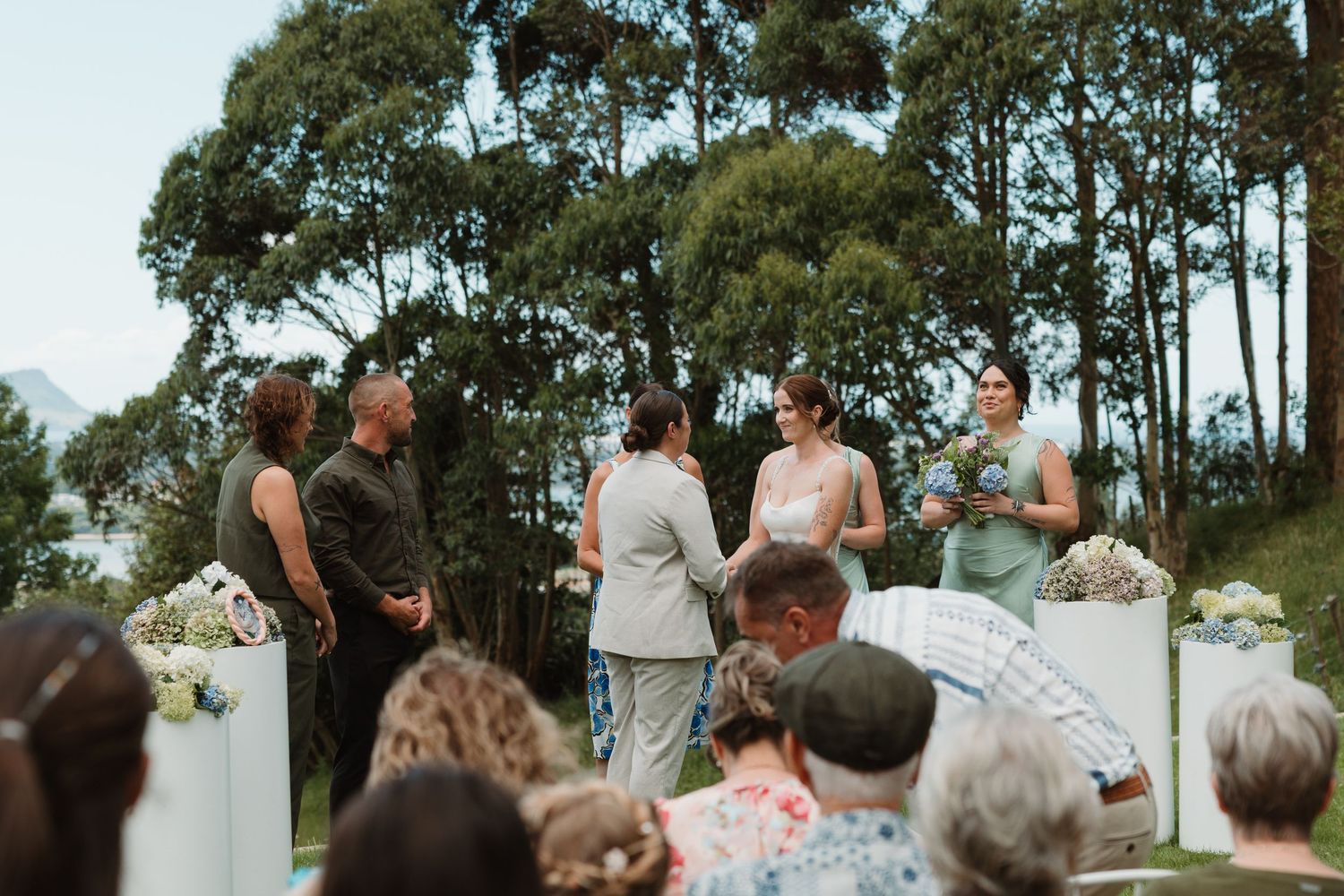 Outdoor wedding ceremony with bridesmaids in sage green dresses, guests seated, surrounded by lush trees.