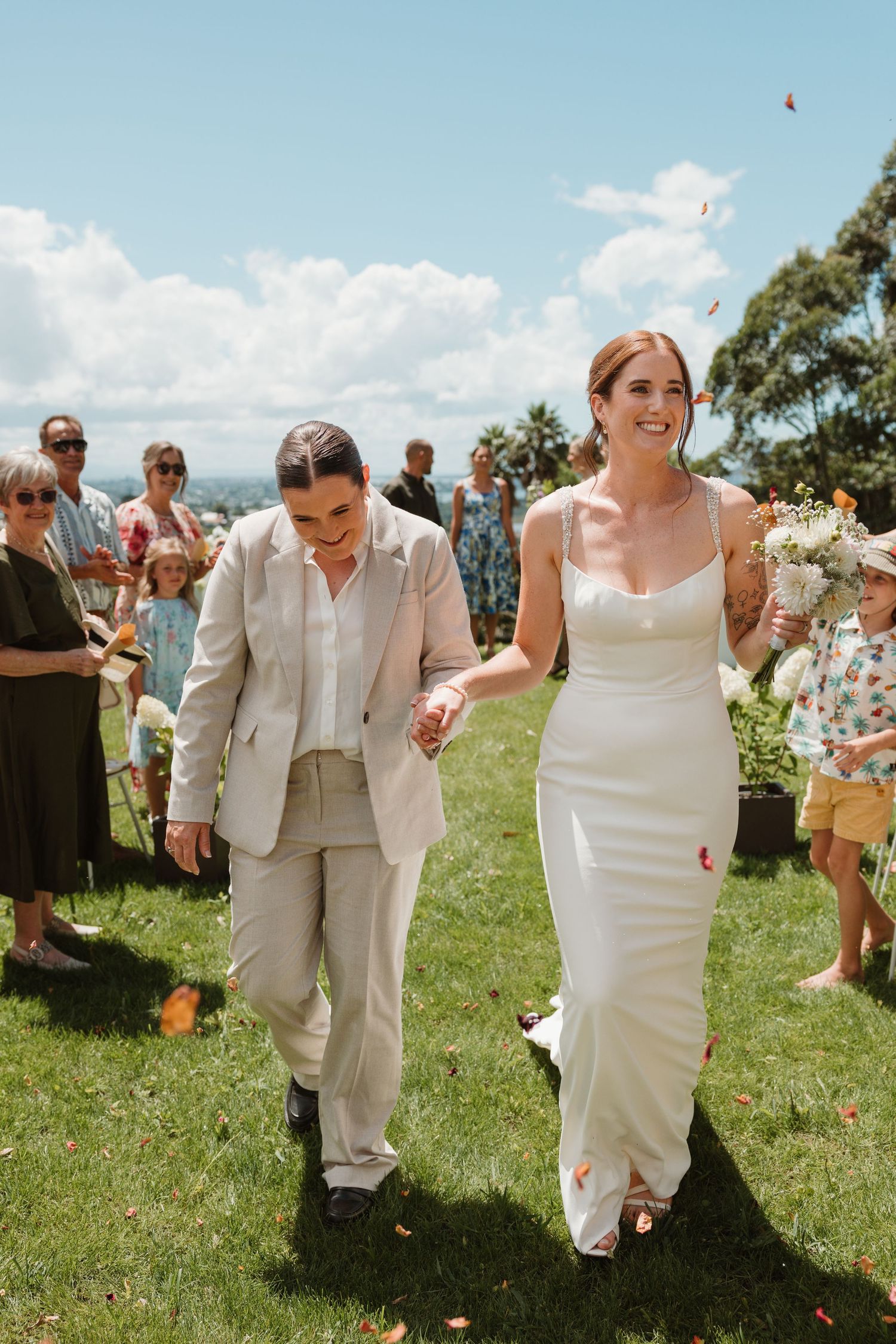 Two brides walk hand in hand down the aisle as guests toss confetti at their outdoor wedding ceremony.