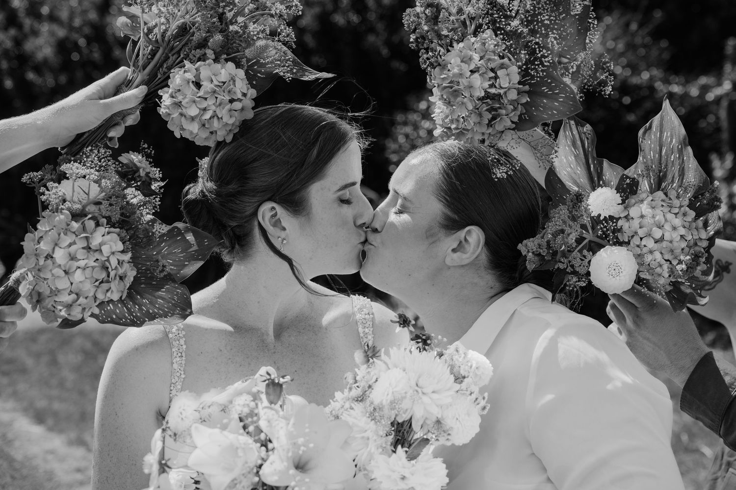 Two brides sharing a kiss surrounded by lush floral arrangements in a black and white wedding photo.