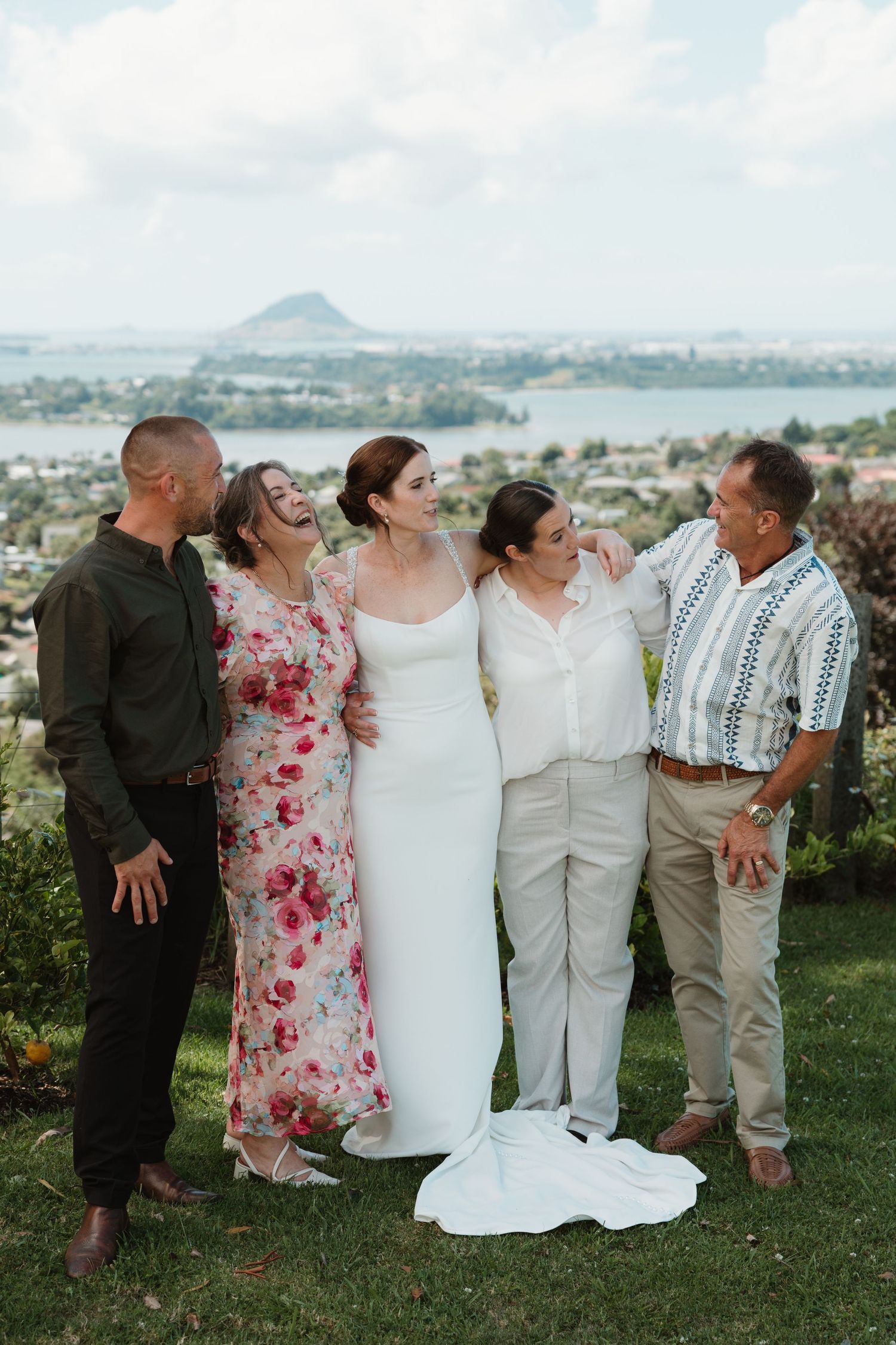 Bride in white gown laughing with family on scenic hilltop overlooking bay and mountain at outdoor wedding celebration.