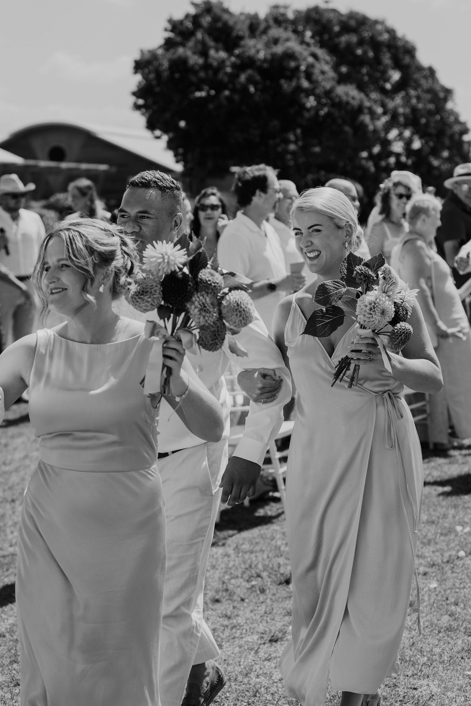 Black and white photo of bridesmaids in elegant dresses holding bouquets, smiling as they walk down the aisle at an outdoor wedding.