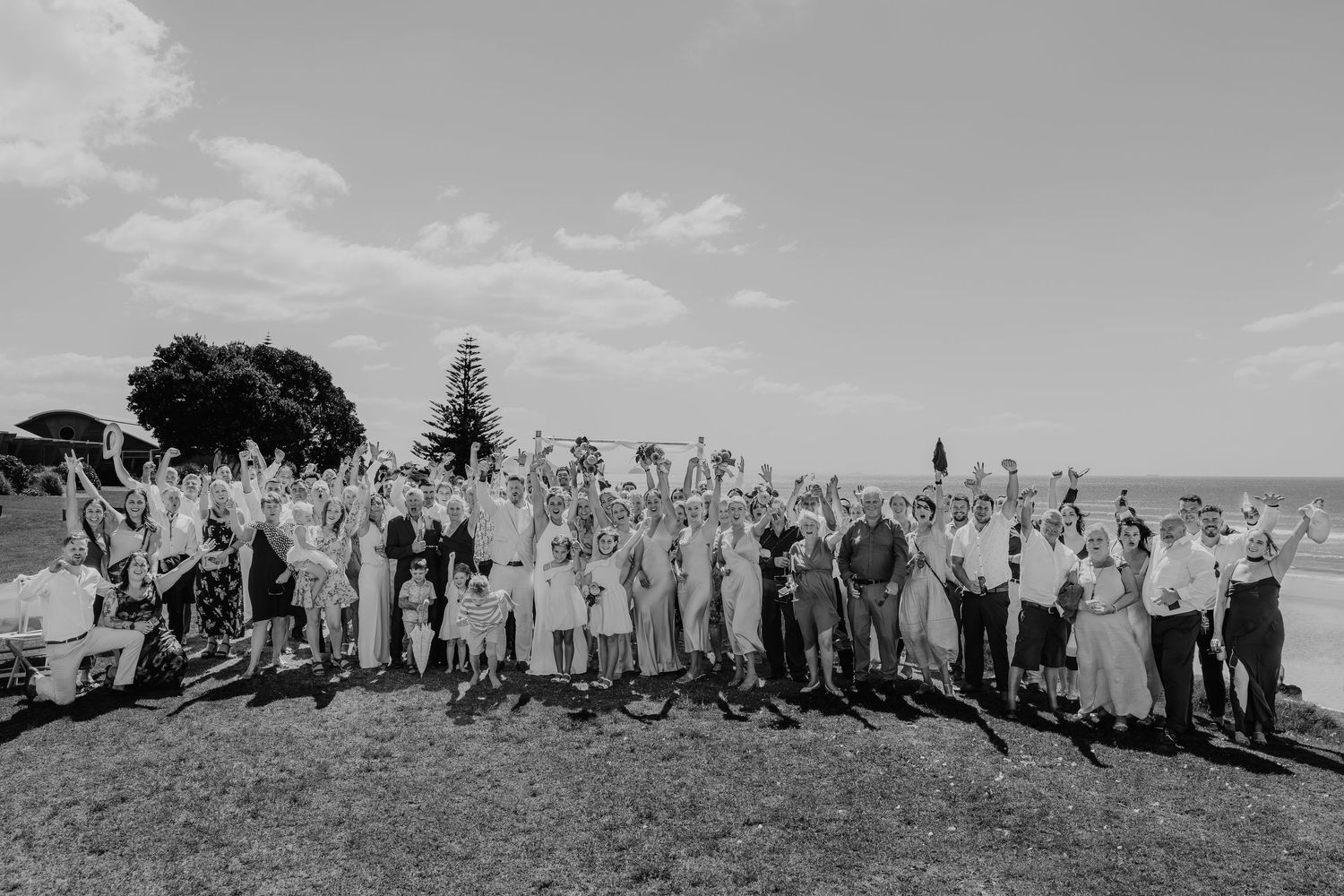 Black and white photo of a large wedding group celebrating outdoors, guests cheering with arms raised under a bright sky.