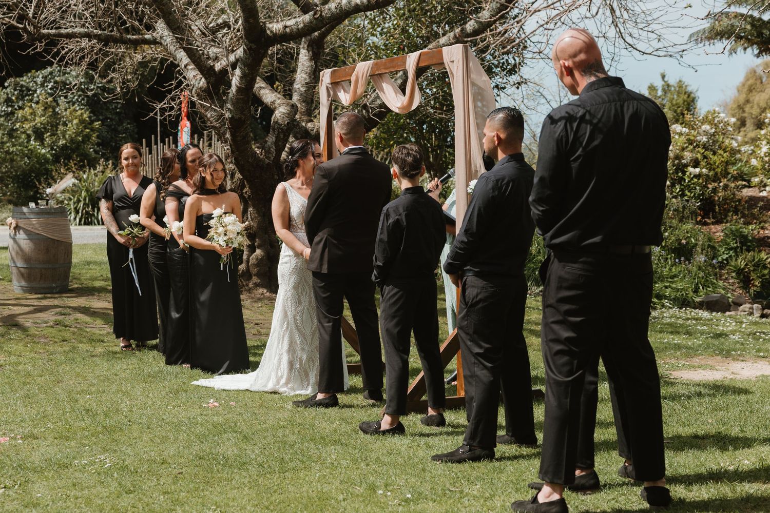 Outdoor wedding ceremony with guests seated on white chairs in a lush garden setting under sunlit trees.