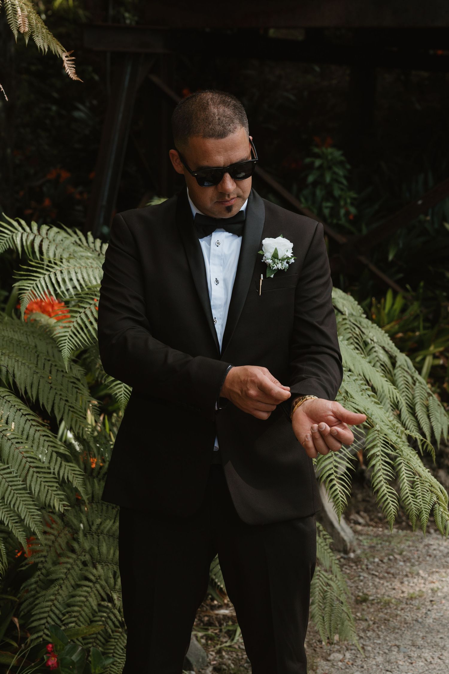 A person in a black tuxedo and sunglasses adjusts their cufflinks while standing near tropical foliage.