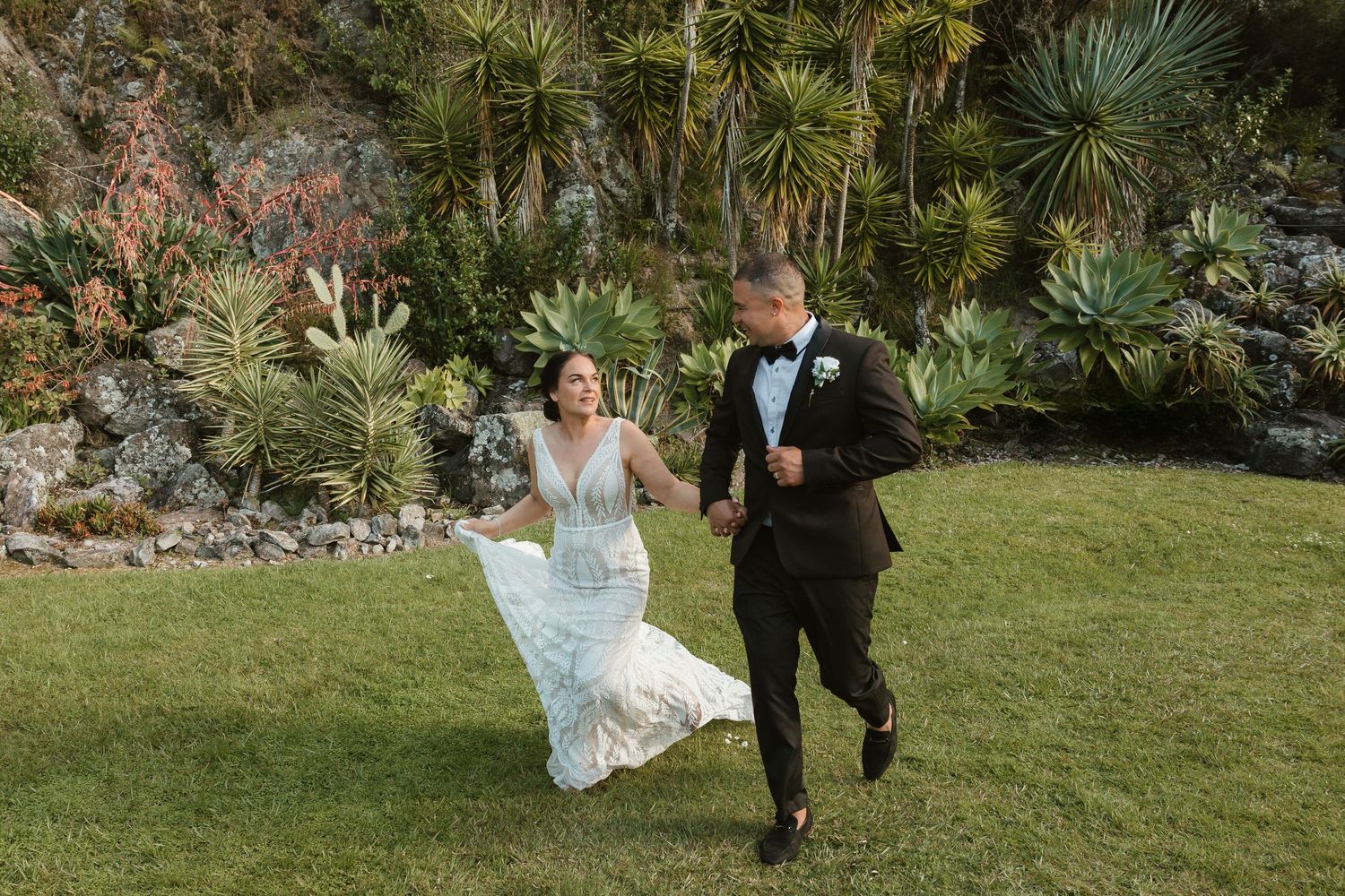 A couple in wedding attire walks together through a lush garden setting during their special day. Bride and groom portraits in Tauranga