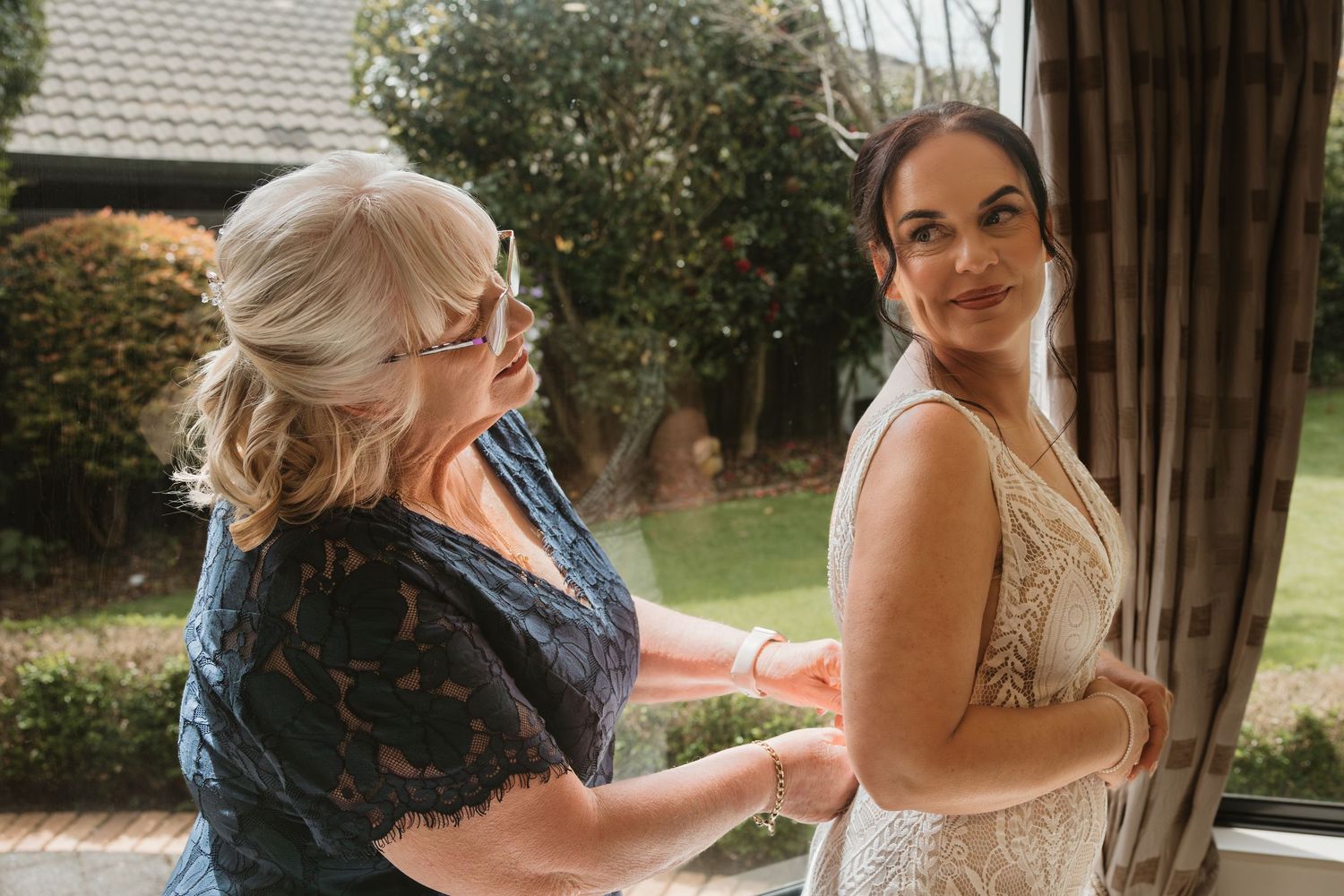 Two people getting ready for a wedding ceremony, with one helping adjust a white lace dress in warm sunlight.