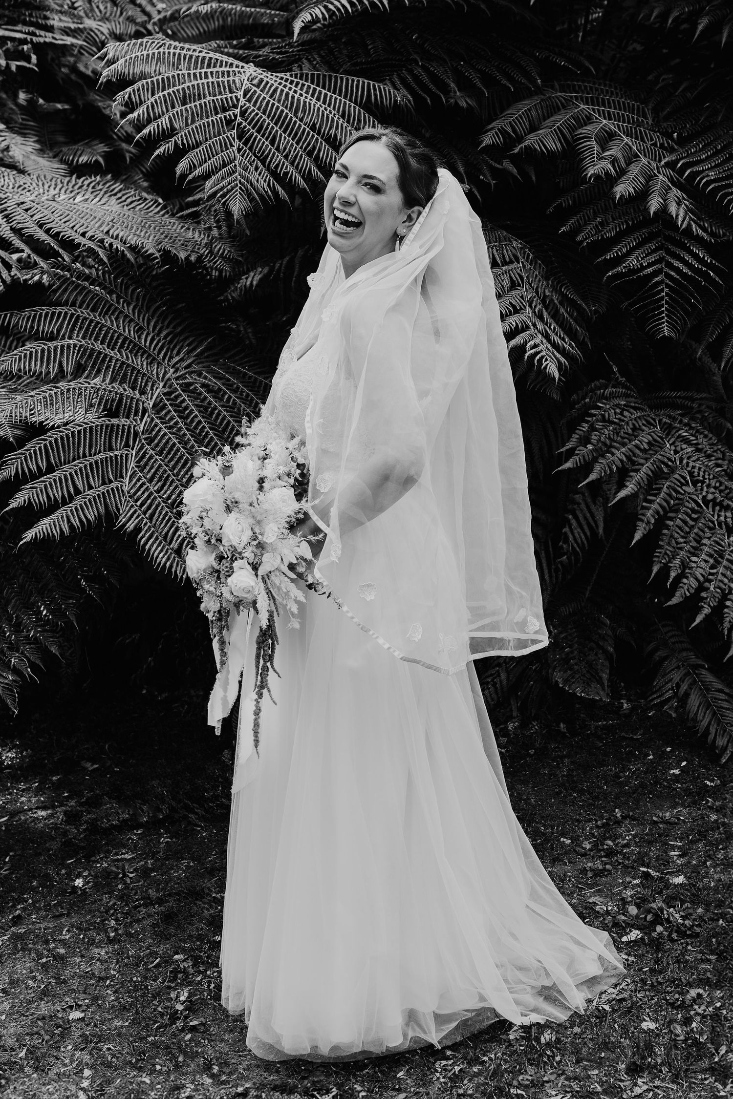 A joyful bride in a classic white wedding gown and veil laughs while holding a bouquet against fern foliage background.