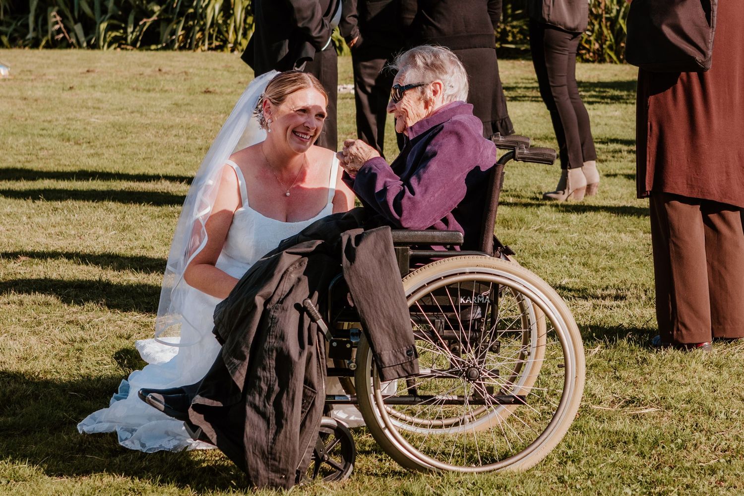 A person in a wedding dress kneels next to someone seated in a wheelchair on a sunny lawn during an outdoor ceremony.