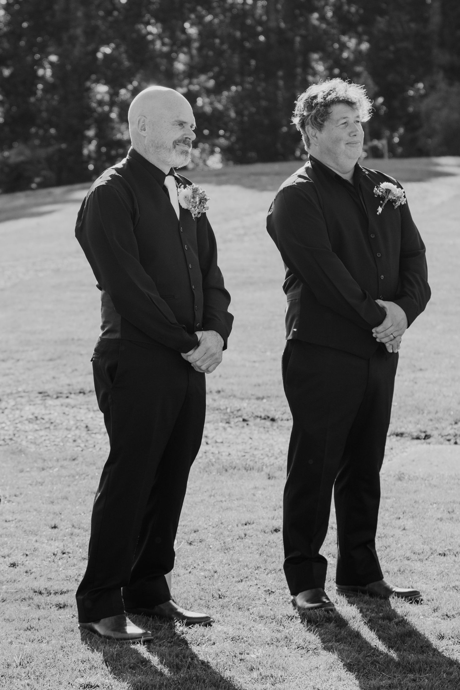 Black and white photo of two people in dark formal attire standing outdoors with boutonnieres on a sunny day.