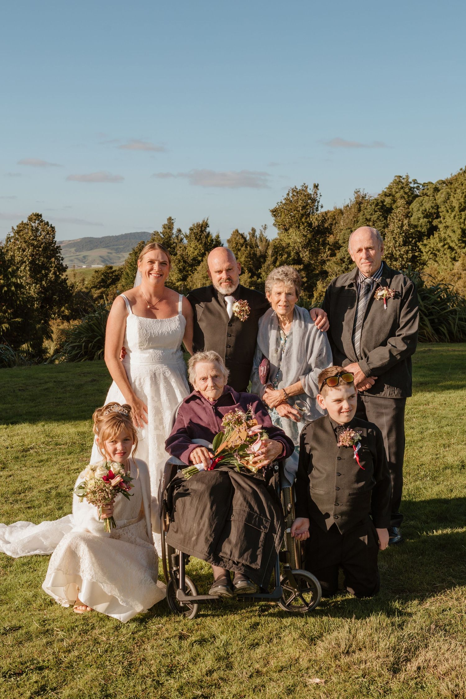 A multi-generational family poses outdoors on a grassy hill with mountains and trees in the background at sunset.