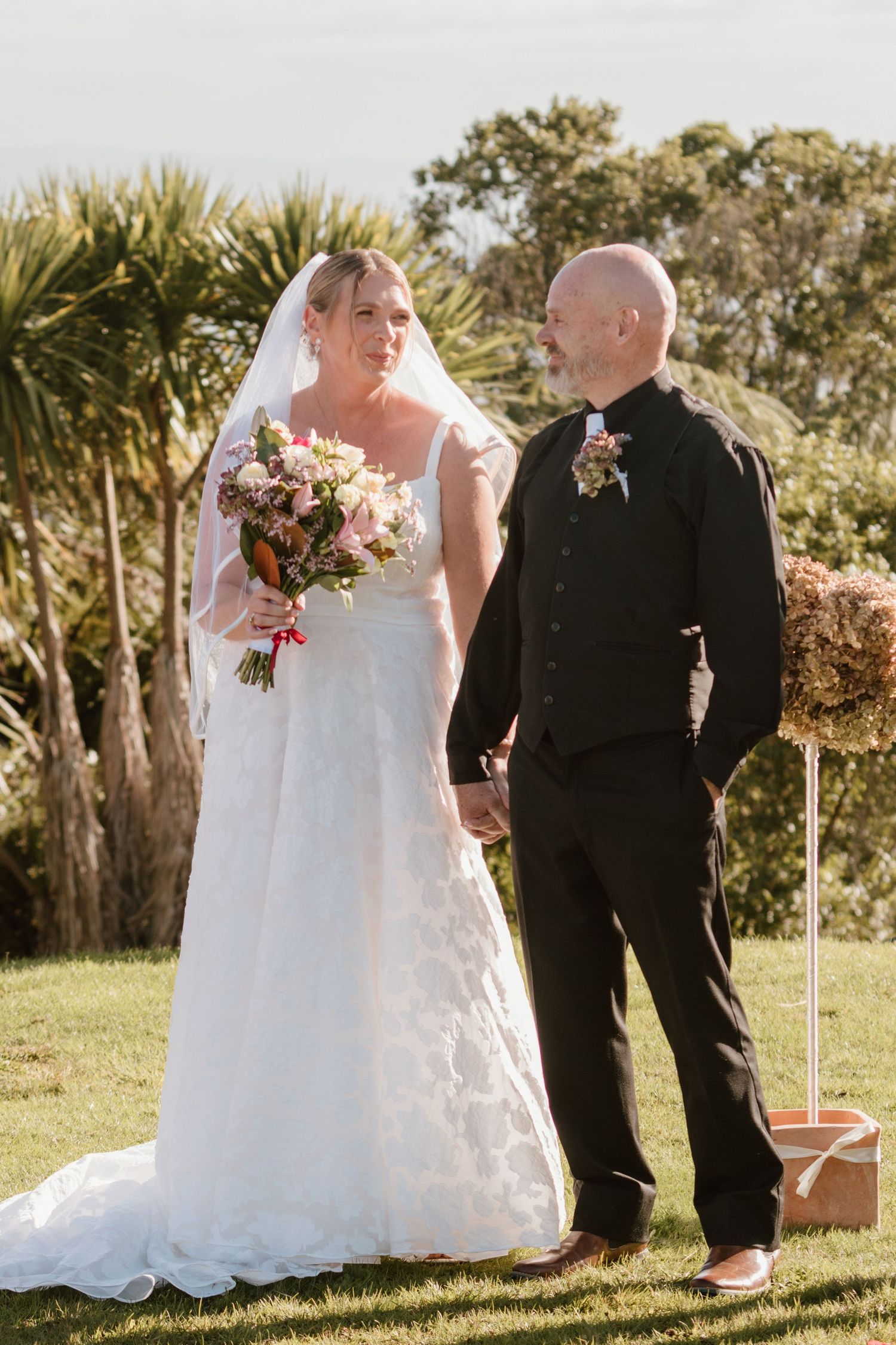 A couple stands together outdoors at their wedding ceremony with palm trees and greenery in the background.