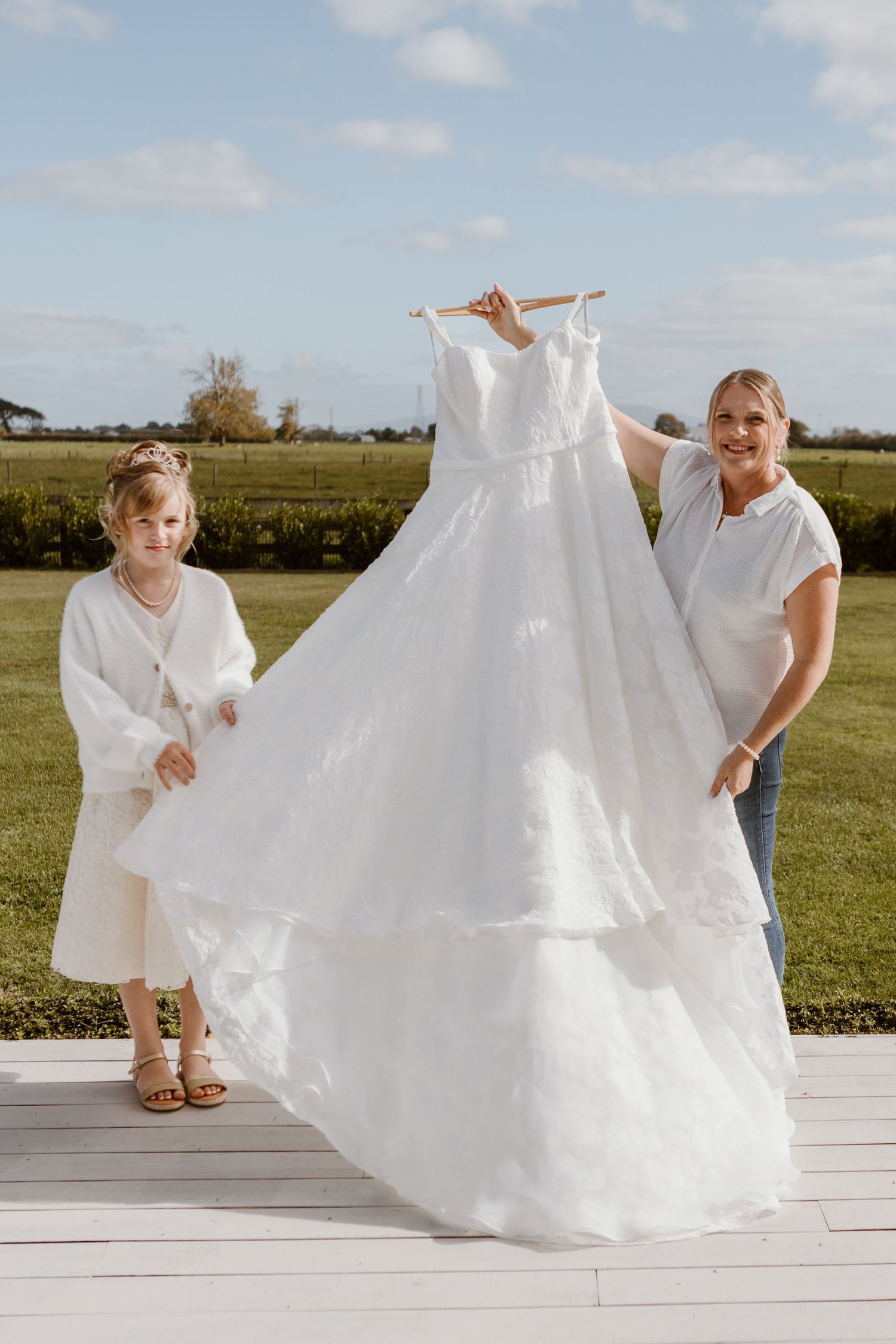 A beautiful white wedding dress is displayed outdoors on a sunny day on a wooden deck with people holding it up for viewing.