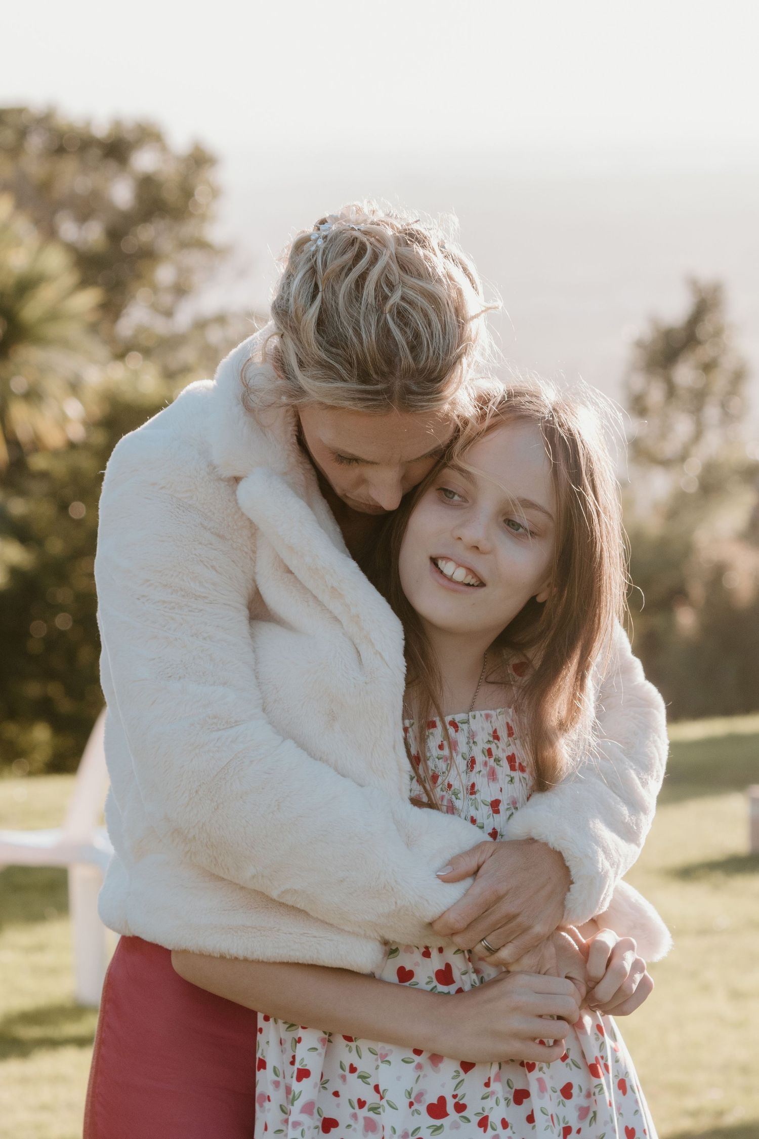 A romantic embrace in sunny nature, with white furry jacket and floral dress creating a tender outdoor moment.