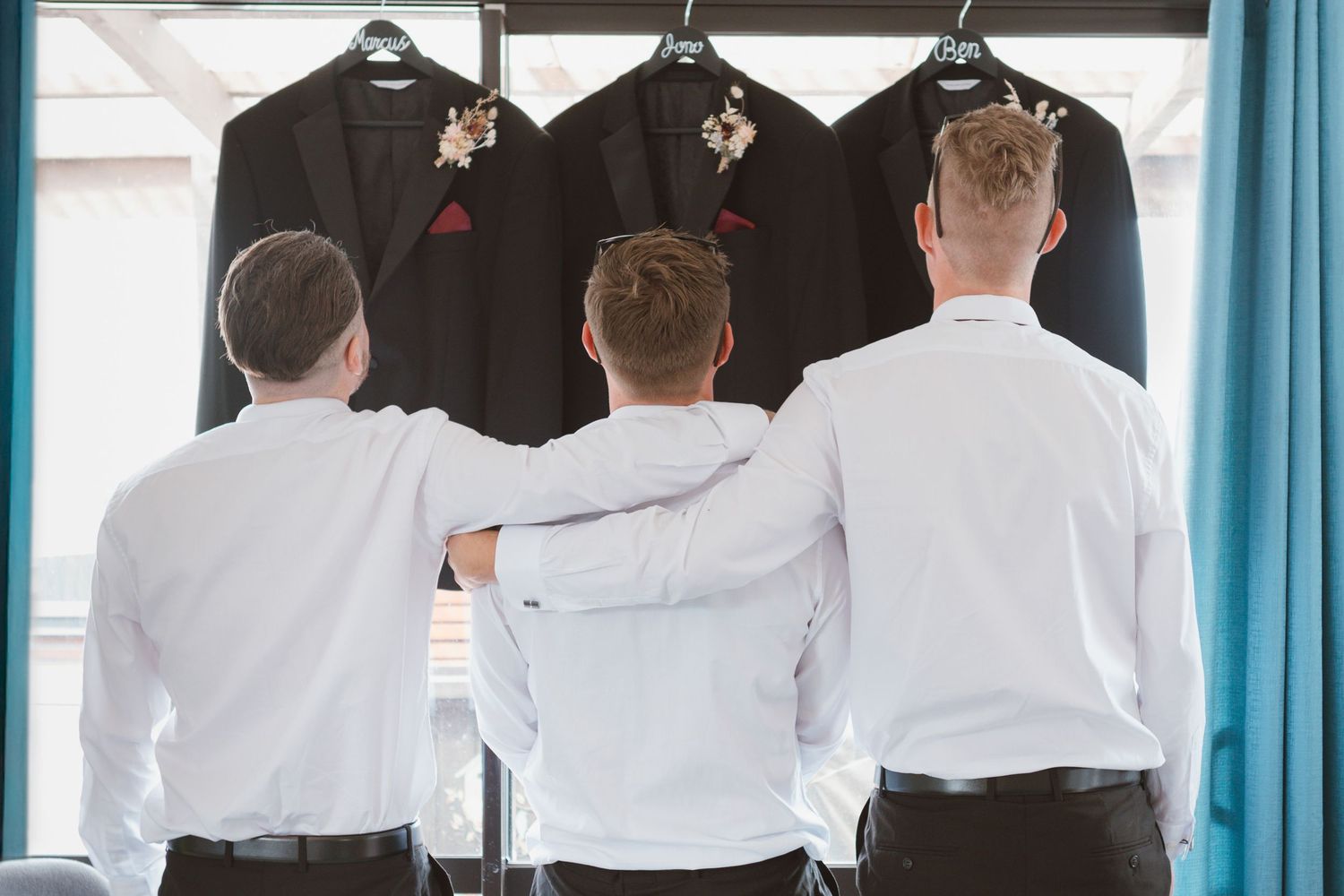 Three groomsmen in white shirts looking at hanging tuxedos before wedding ceremony.